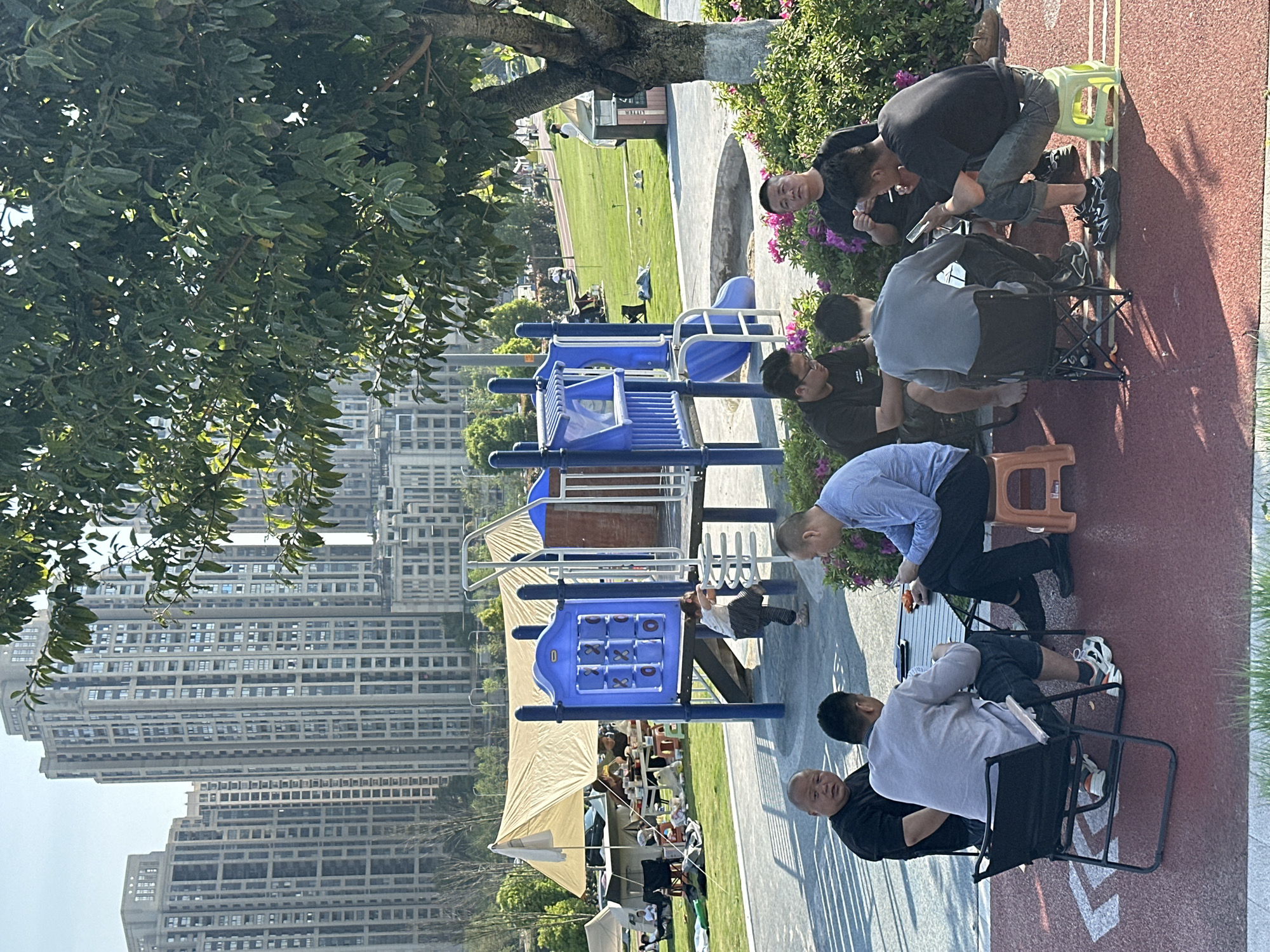 The image depicts a park scene with high-rise buildings in the background, suggesting an urban environment. A group of people are gathered near a children's playground, possibly engaged in a social activity. Some are seated on chairs and stools, while others are standing. The playground includes a climbing structure and a tic-tac-toe board. A tent is visible in the distance, indicating a public gathering or event. The location is likely Jiangjin, China, based on visual characteristics of the environment.