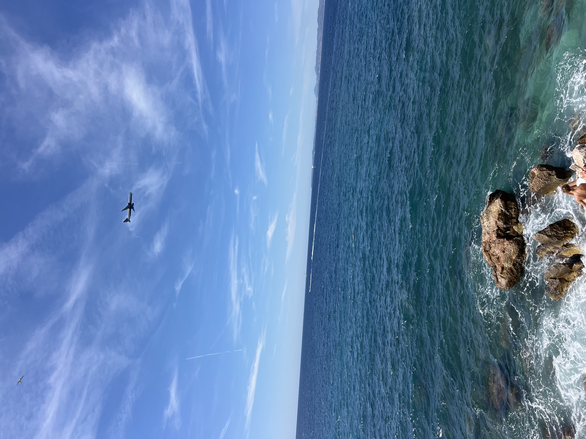 The image depicts a coastal scene with a vibrant blue sky and the Mediterranean Sea. In the sky, a white airplane is visible flying from left to right, accompanied by scattered white clouds and a contrail from another aircraft. Two birds are also present in the sky. The sea is a deep blue, with a few whitecaps indicating gentle waves breaking against a rocky shore in the foreground. Several boats are visible in the distance on the water. On the rocks in the lower part of the image, two individuals are seated, appearing to be enjoying the view. The location is likely along the coastline of Nice, France, given the characteristic clear blue waters and rocky shoreline.