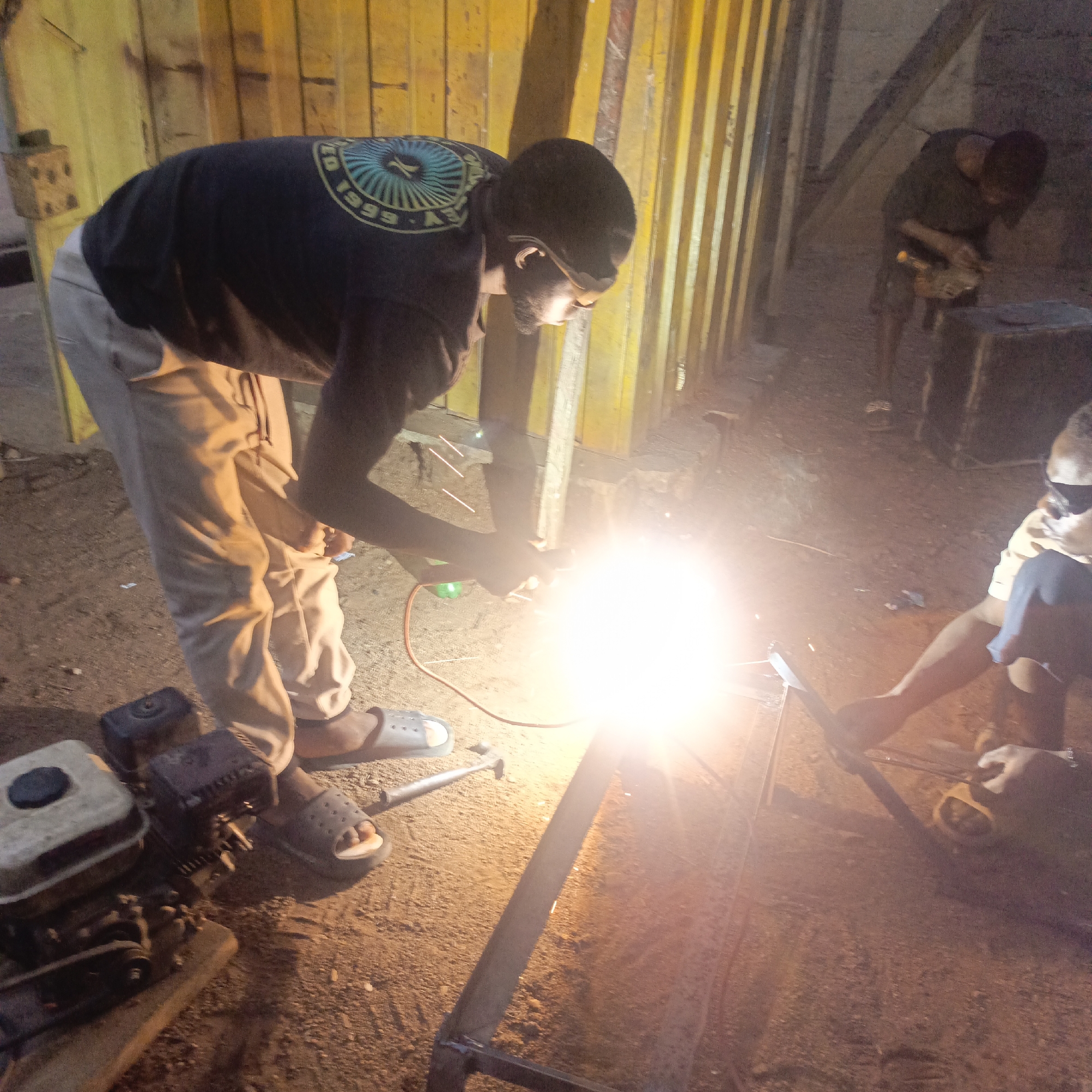 Akwanga, Nigeria. A person wearing a black t-shirt with a circular design, light-colored pants, and sandals welds metal in an outdoor workspace with a dirt floor. Another person, wearing dark clothing, works in the background, possibly also welding. Additional individuals sit nearby. A hammer and a gas-powered generator sit on the ground near the welder. The background includes a yellow metal wall.