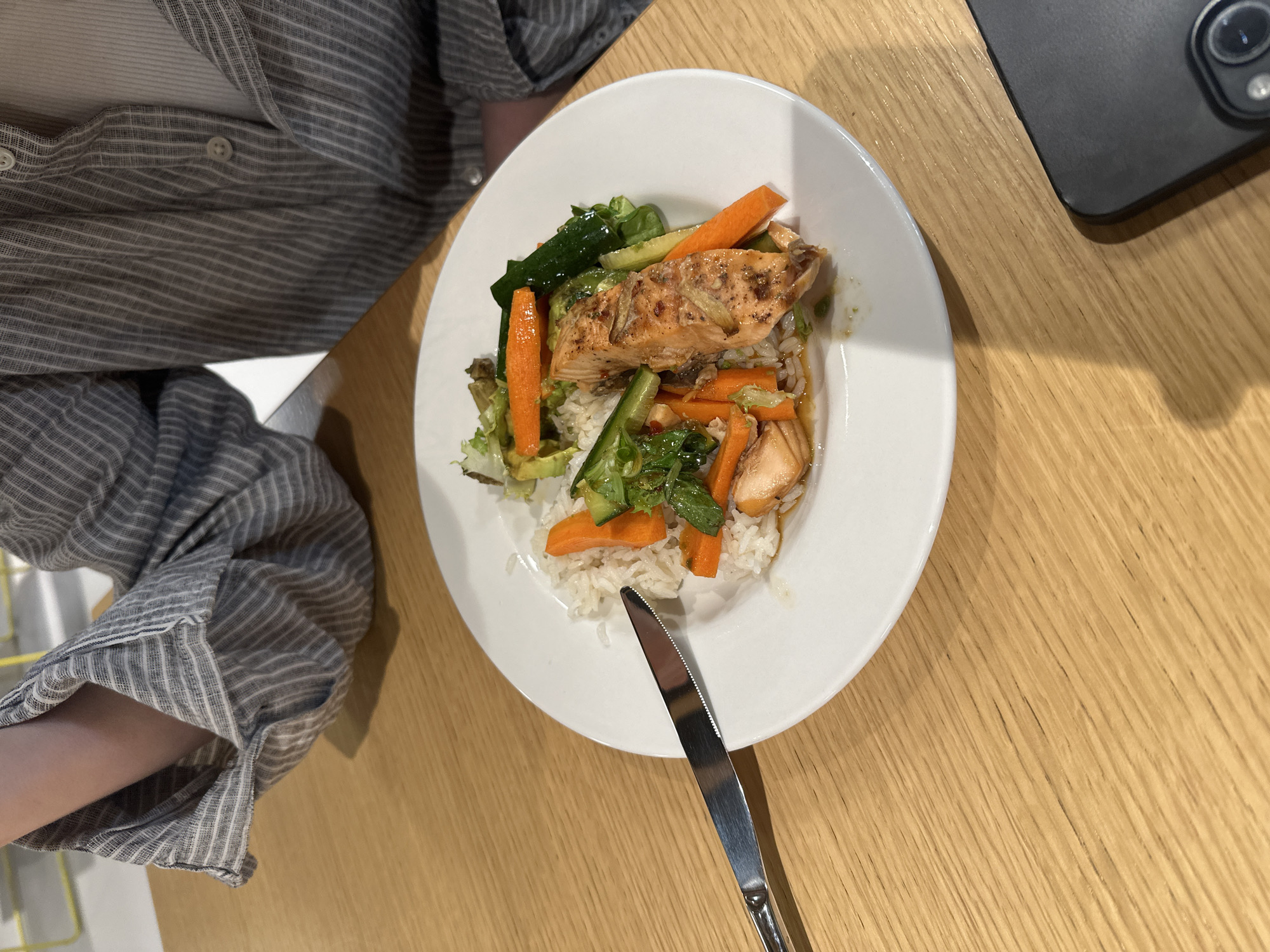 A top-down shot shows a white plate of food consisting of rice, salmon, carrots, zucchini, and greens on a light wood table. A silver knife rests on the edge of the plate. Part of a person is visible to the left, wearing a striped shirt. A black phone sits in the top right corner of the image. No information suggests this image was taken in London, United Kingdom.