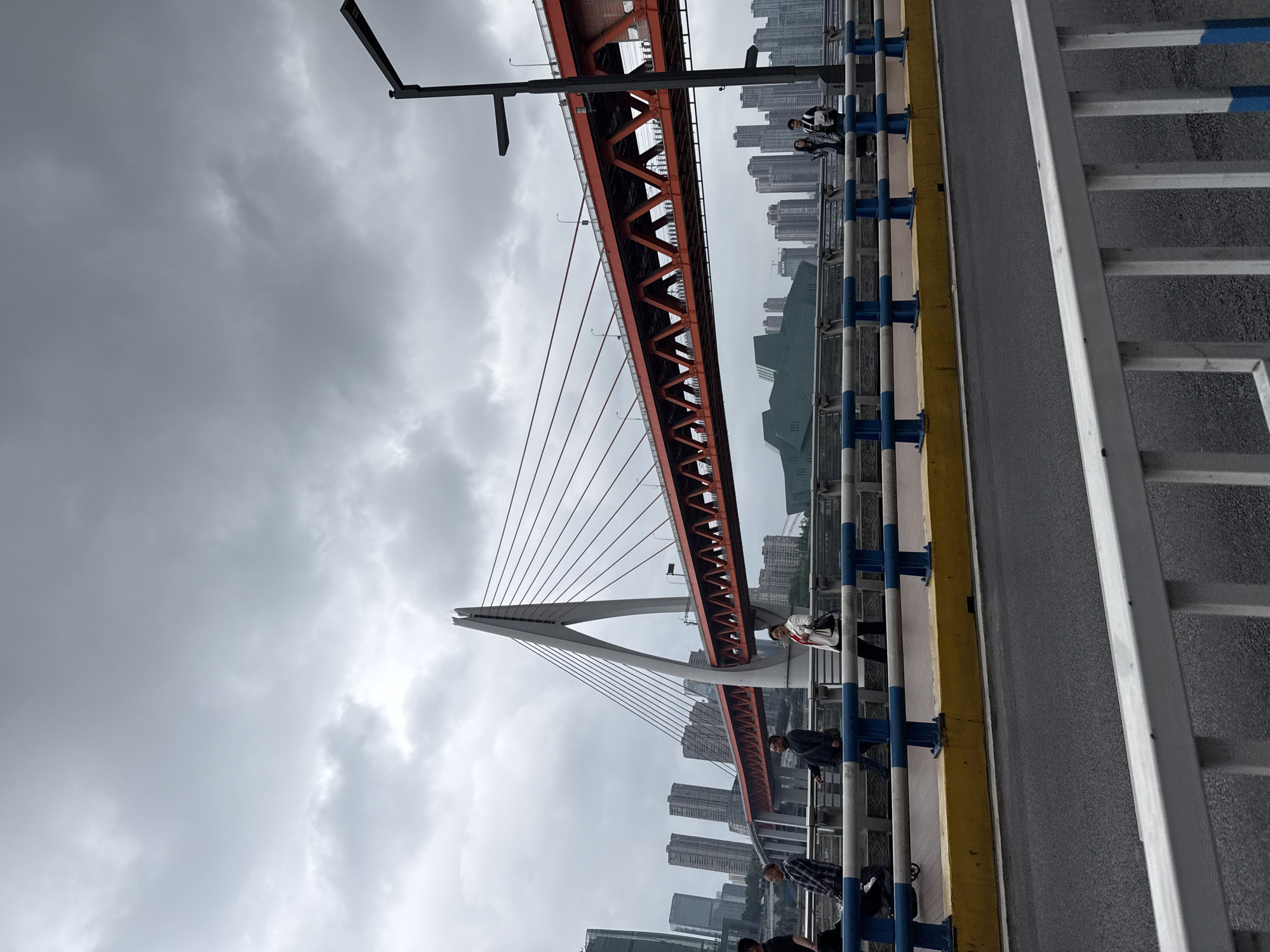The image captures a panoramic view of an urban landscape, centered around a prominent red cable-stayed bridge under an overcast sky. The photograph is taken from an elevated pedestrian walkway, likely part of the bridge itself, looking towards the main span and the city beyond.
In the foreground, the grey asphalt of the walkway is visible, bordered by a yellow curb on one side and blue and white metal railings on the other. Several individuals are present on this walkway. On the right side, a person in a white top and dark pants is standing by the railing, possibly looking out at the view. Further down the walkway, other people are visible, including one in a plaid shirt and another in a dark jacket, also near the railings or walking.
The main subject is a large red bridge, characterized by its intricate lattice-like truss structure and multiple cables extending from a tall, white, A-frame pylon. The cables suspend the bridge deck, which is also reddish-orange. This bridge stretches across what appears to be a river or valley, connecting different parts of the city. Given the location context, this is likely the Dongshuimen Bridge in Chongqing.
In the background, a dense cluster of tall buildings and skyscrapers defines the city skyline, indicative of a large metropolitan area. The sky is heavily clouded and grey, suggesting an overcast day with diffused light. A street light fixture is partially visible in the upper left corner of the frame. There is no visible text in the image. The overall impression is one of a bustling city scene observed from a major infrastructural landmark on a cloudy day.