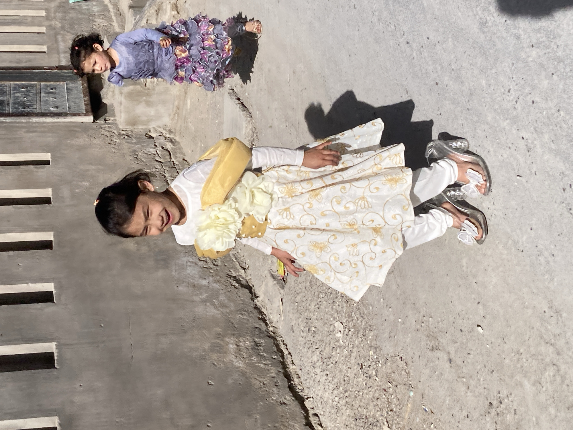 Two young girls are on an uneven outdoor concrete surface near a building in Quetta City Tehsil, Pakistan. One girl wears a light-colored dress with gold-colored floral designs and white long sleeves. She is adorned with yellow accessories, including a sash and a flower. She wears clear sandals. The other girl wears a blue and purple dress with a ruffled skirt. The background shows a building with a series of dark, rectangular openings.
