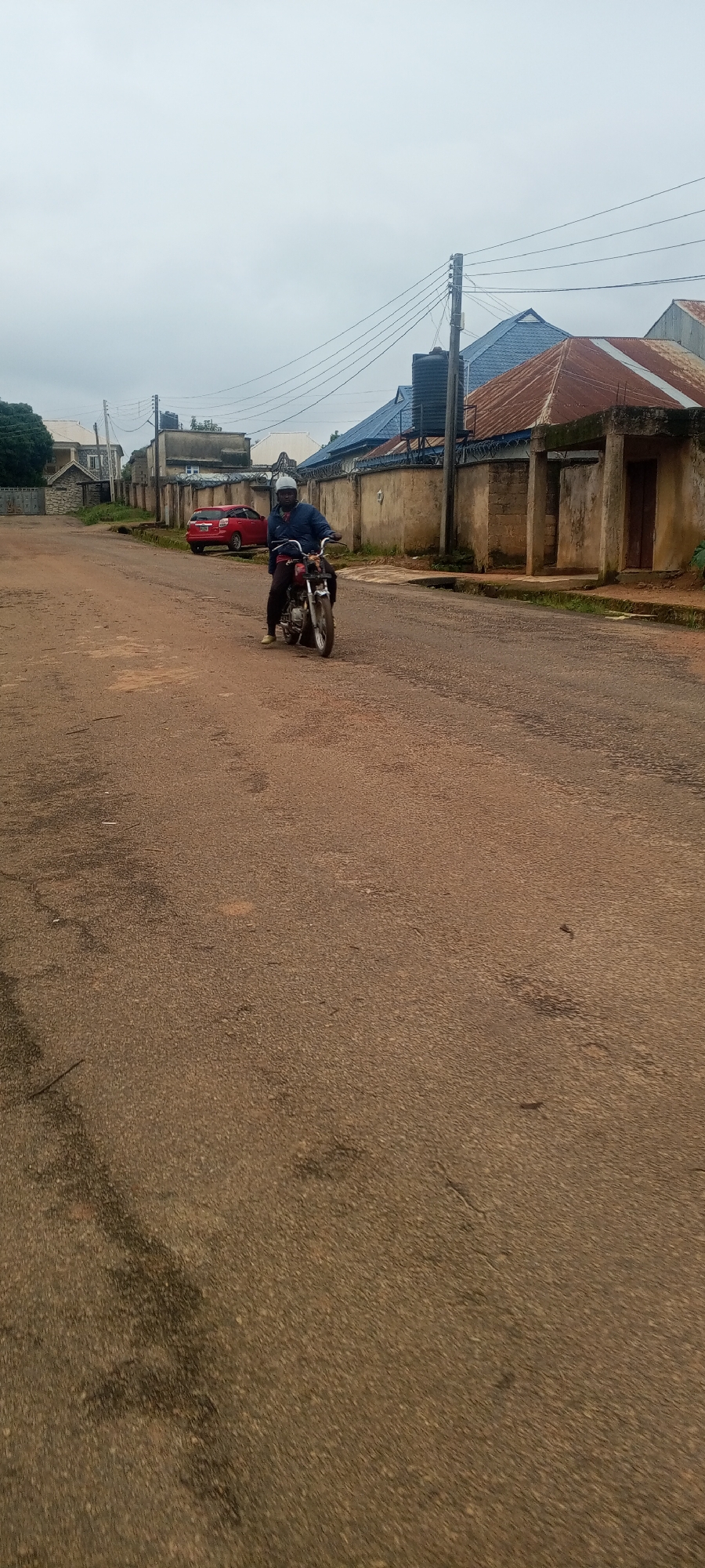A man wearing a blue jacket, dark pants, and a white helmet is riding a red motorcycle on a paved road. The motorcycle has a silver exhaust pipe. The man is looking forward and appears to be in motion. The road is a reddish-brown color and shows signs of wear and tear. In the background, there are several buildings with various roof styles, including corrugated metal and blue roofing. Power lines are visible crossing the sky, supported by utility poles. A red car is parked on the left side of the road. The sky is overcast and cloudy. The scene is likely a street in Bahit, Nigeria.