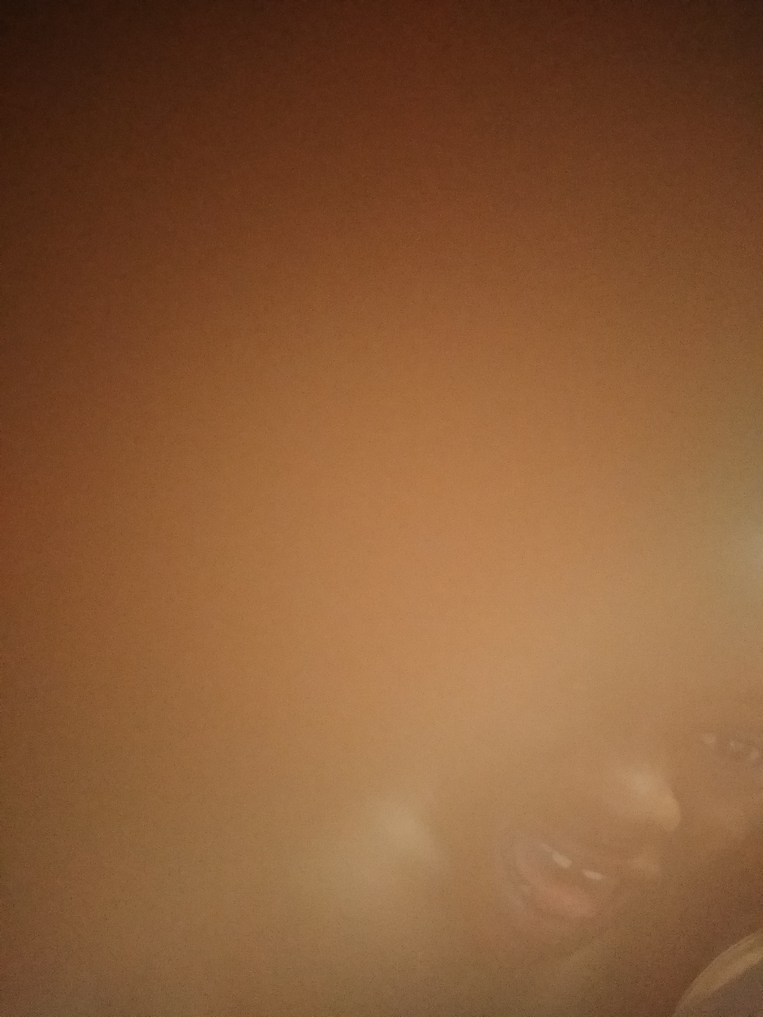 The image presents a highly blurred, close-up view dominated by warm, reddish-brown tones. The overall scene appears underexposed or taken in low light conditions, contributing to significant blur and image noise.
In the bottom right quadrant, a very indistinct and out-of-focus partial human face is visible. Key features that can be discerned include what appears to be an open mouth, possibly revealing teeth, suggesting an expression of a wide smile or laughter. Above the mouth, blurry shapes hint at a nose and possibly a cheek or eye area, but these are too indistinct for clear identification. The visible skin tone appears dark, blending into the warm background.
A bright, diffused light source or reflection is present along the far right edge of the frame, creating a strong highlight that further obscures details in that area. The background is a solid, warm brown color with subtle gradients, lacking any discernible objects or structural elements. Due to the extreme blur and lack of context, no specific activity, event, or precise location cues are evident within the image itself. The time of day is not determinable, though the warm, dim lighting suggests an indoor setting or artificial light. No visible text is present.