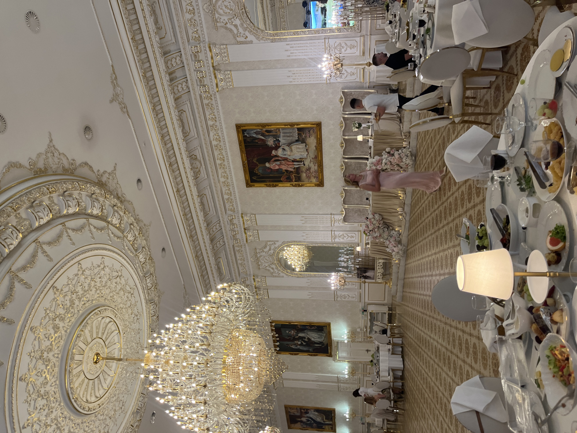 This is a photograph of a richly decorated banquet hall, likely within the city of Dubivtsi, Ukraine. The hall features ornate, gold-trimmed plasterwork on the ceiling and walls, illuminated by a grand, multi-tiered crystal chandelier. Paintings in ornate frames adorn the walls, and large mirrors reflect the opulent decor. Several round dining tables are set with white tablecloths, dinnerware, and glassware, suggesting a catered event. In the foreground, a table is laden with food, including fruit, salads, and what appears to be fried food. People are visible in the hall; a woman in a pink dress stands near a table, conversing with a man carrying a tray. Other guests are seated at tables or mingling in the background. The overall impression is one of a formal and lavish celebration.