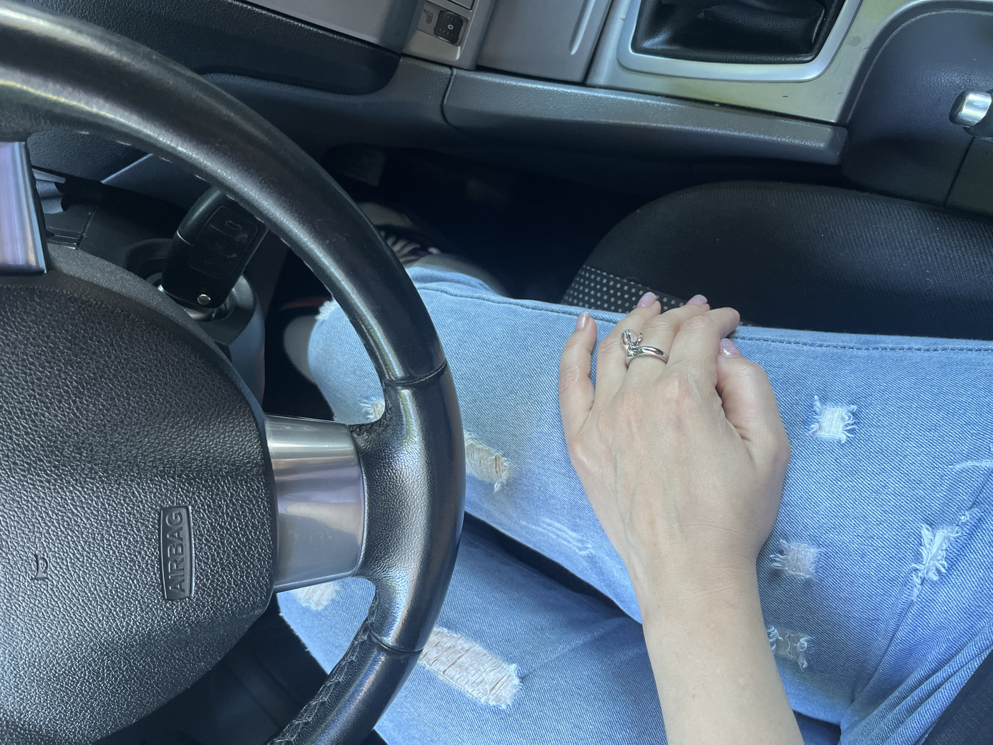 The image shows a first-person perspective from inside a car. The subjects are a person's hand and lap, dressed in distressed light-wash denim jeans. The hand is adorned with a silver ring that appears to be shaped like a bow or knot. The setting is the interior of a car, with a black steering wheel prominently featured on the left. The steering wheel has a silver accent and an "AIRBAG" label. In the background, part of the car's dashboard and console are visible, including what appears to be a gear shift lever. A car key is inserted into the ignition. The lighting suggests it is daytime, with natural light entering the car. There are no specific cues to indicate weather or a particular emotion. The location context is Voronezh, Russia.