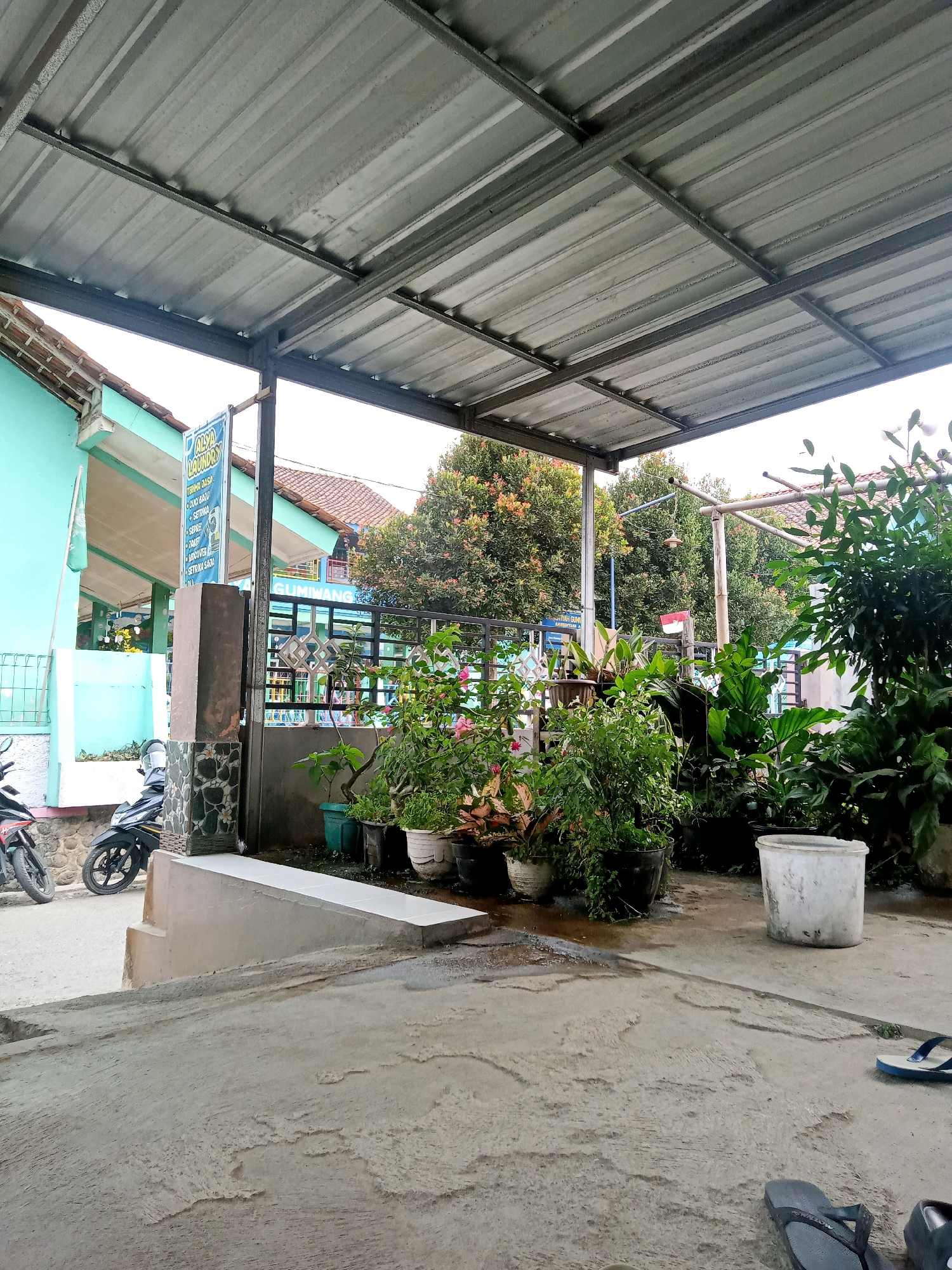 This is a ground-level, outdoor, daytime, vertical shot of an area under a metal canopy in Gumiwang, Indonesia. The canopy's corrugated metal roof is supported by metal beams, and its underside is visible. A light blue building with a tile roof is on the left, with a white flag flying from a pole. A sign with text in blue and white is attached to a pillar. A dark-colored pillar with mosaic tile accents is in the foreground. Two motorcycles are parked on the left side of the frame. A paved ramp leads up from a concrete ground. Several potted plants are arranged on the right side, in front of a metal fence. A large white bucket and a pair of blue flip-flops are on the concrete ground to the right. The background shows trees and other buildings. The ground is concrete, with some areas appearing wet.
