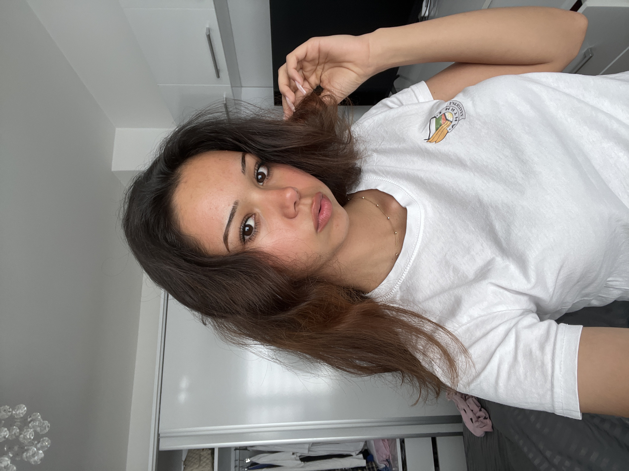 In this medium shot, a young woman with long, dark brown hair is positioned centrally, facing slightly to her left. She wears a white t-shirt with a small, colorful emblem on the left chest. The emblem depicts a shield with a coat of arms, featuring a stylized plant and what appears to be wheat or barley stalks. Above the shield are the words "АНТИМ" and below it, "ЗЛАТОГРАДЪ," indicating the city of Zlatograd, Bulgaria. Her right hand is raised, touching a strand of her hair. The background shows a cluttered room with a wardrobe on the left, containing clothing on hangers and shelves. To the right, white cabinets with silver handles are visible, along with a dark electronic device, possibly a television or computer monitor. The room is illuminated by ambient lighting, creating a soft, diffused glow.