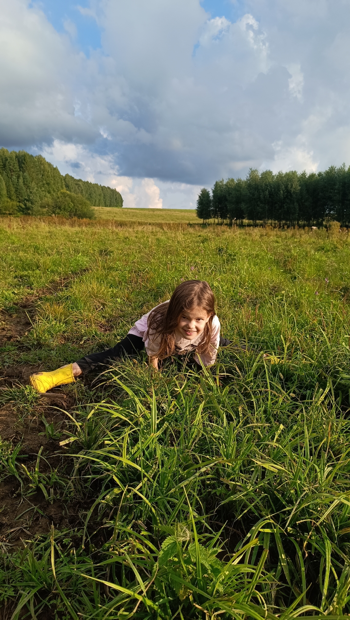 A young girl with long brown hair is lying on her stomach in a grassy field, looking up at the camera with a smile. She is wearing a light pink long-sleeved shirt, black pants, and bright yellow rubber boots. The grass around her is tall and green, with some patches of dark, wet earth. In the background, there is a line of trees that stretches across the horizon, with a field of grass in between. The sky above is filled with dramatic clouds, with patches of blue sky showing through. The scene appears to be set in the late afternoon, with the sun casting long shadows. The overall mood is joyful and playful. The location is Karsha, Russia.