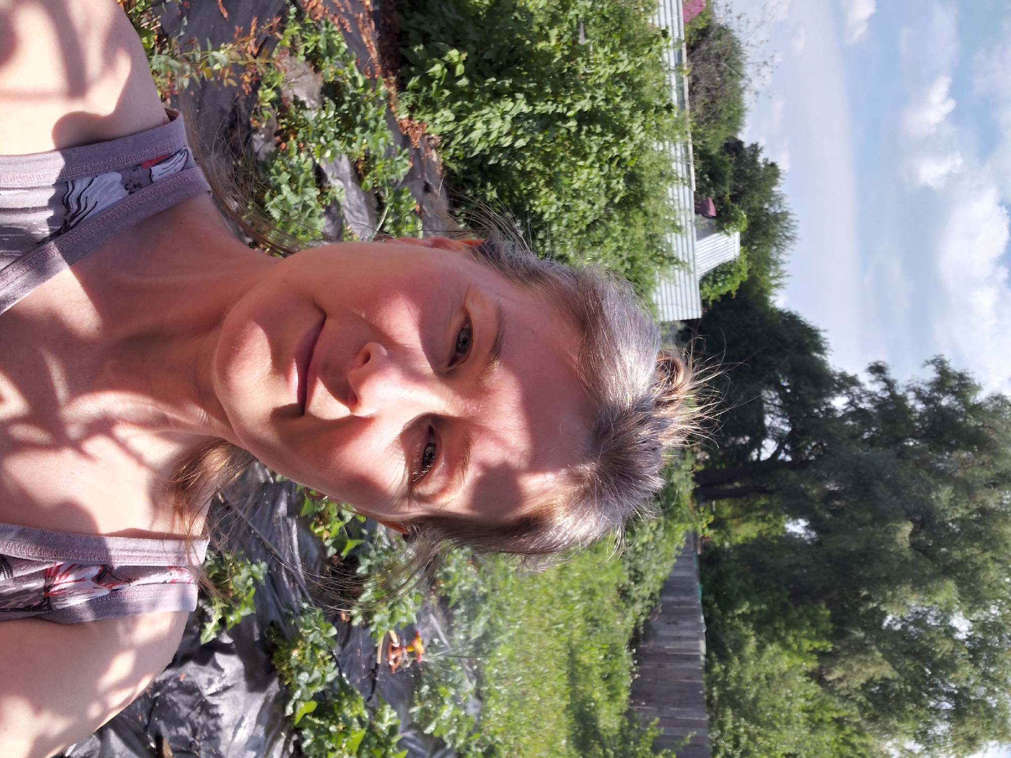 A woman with fair skin, light brown hair pulled back, and gray eyes smiles slightly as she takes a selfie outdoors in a garden. She is wearing a sleeveless top with a patterned design. Behind her, rows of plants, possibly strawberries, are covered with black plastic sheeting. Further back, there are dense green bushes, a wooden fence, and trees under a partly cloudy sky. The scene appears to be a suburban or rural garden setting, consistent with locations in the Novosibirsk Oblast region of Russia.