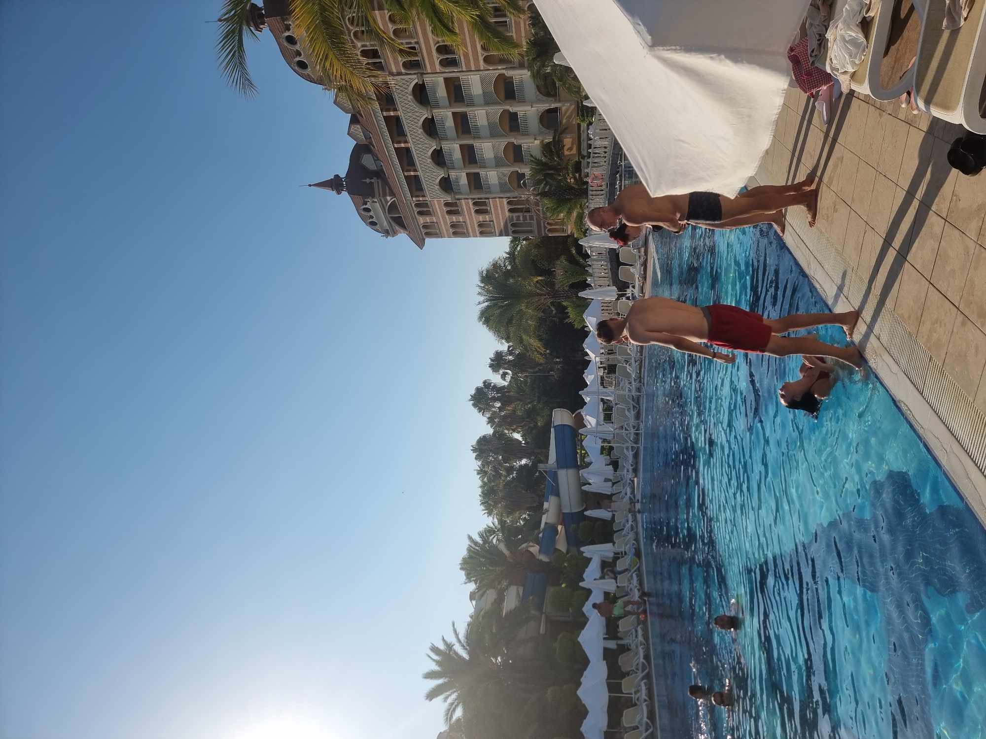 The image depicts a poolside scene at a resort in Manavgat, Turkey. In the foreground, four individuals are visible. Two young men stand on the tiled edge of a swimming pool, one in red shorts and the other in dark swim trunks. To their right, a woman with red hair wearing a black bikini top and another man in dark swim trunks also stand by the pool. The swimming pool is filled with clear blue water, and several people are swimming or wading in it. In the background, rows of white lounge chairs and umbrellas are arranged, indicating a resort setting. Beyond the pool area, lush green palm trees and other tropical foliage are abundant. A large, multi-story hotel with ornate architecture, featuring arched windows and decorative balconies, dominates the far background. A water slide complex is also visible amongst the trees. The sky is clear and bright blue, with the sun casting long shadows across the pool deck. The overall impression is one of a leisure resort on a sunny day.