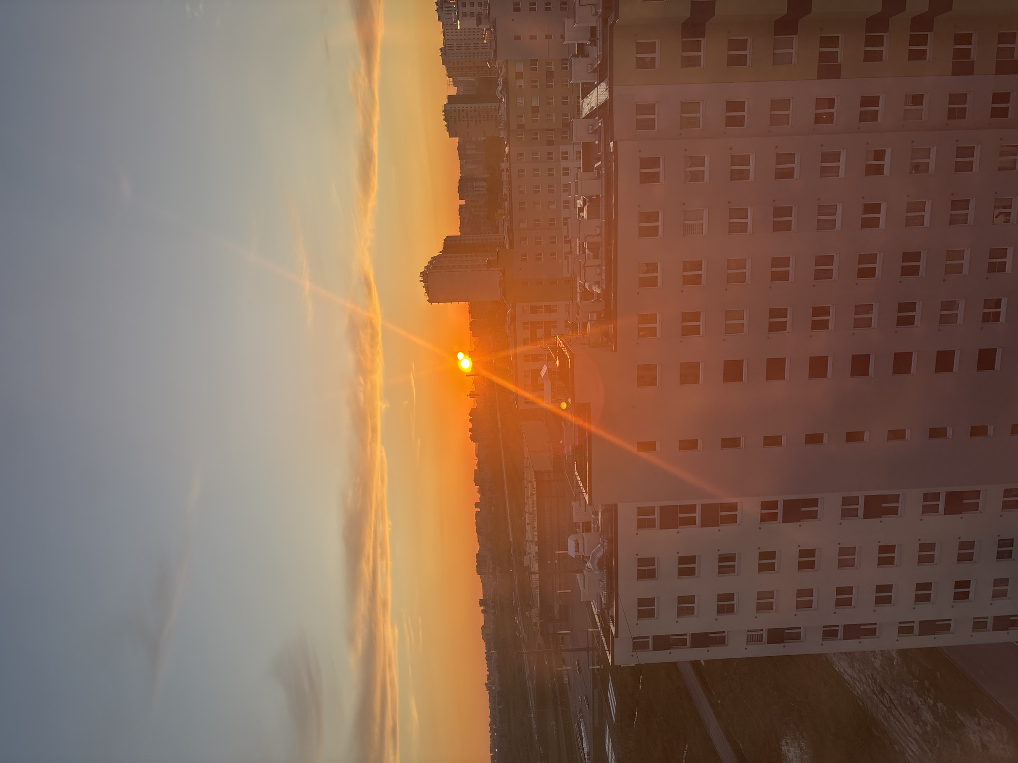 The image depicts a sunset in an urban environment. The sun is low on the horizon, partially obscured by clouds, casting a warm orange glow across the sky and buildings. Several high-rise buildings are visible in the background, with a large, multi-story building dominating the foreground on the right side of the image. The building's facade is marked by numerous windows arranged in a grid-like pattern. Below the building, there appears to be a road or ground-level area. The general location is in the city of Unknown, Unknown.