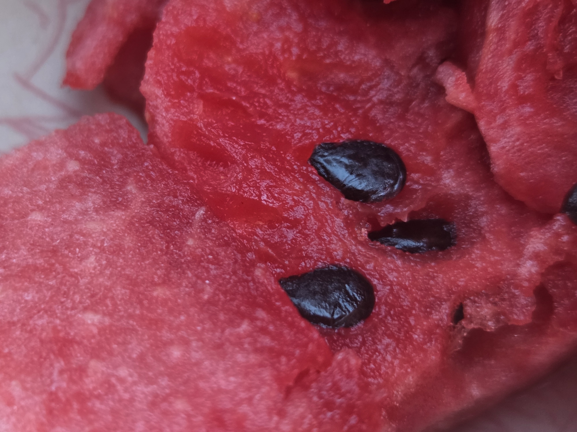 A close-up, macro photograph captures the textured red flesh of a watermelon with several black seeds. Two seeds are positioned to resemble eyes, and a smaller seed or piece of flesh below them forms a mouth, creating a skull-like face. The background is out of focus and appears to be a light-colored surface. No individuals or specific locations are identifiable in the image.