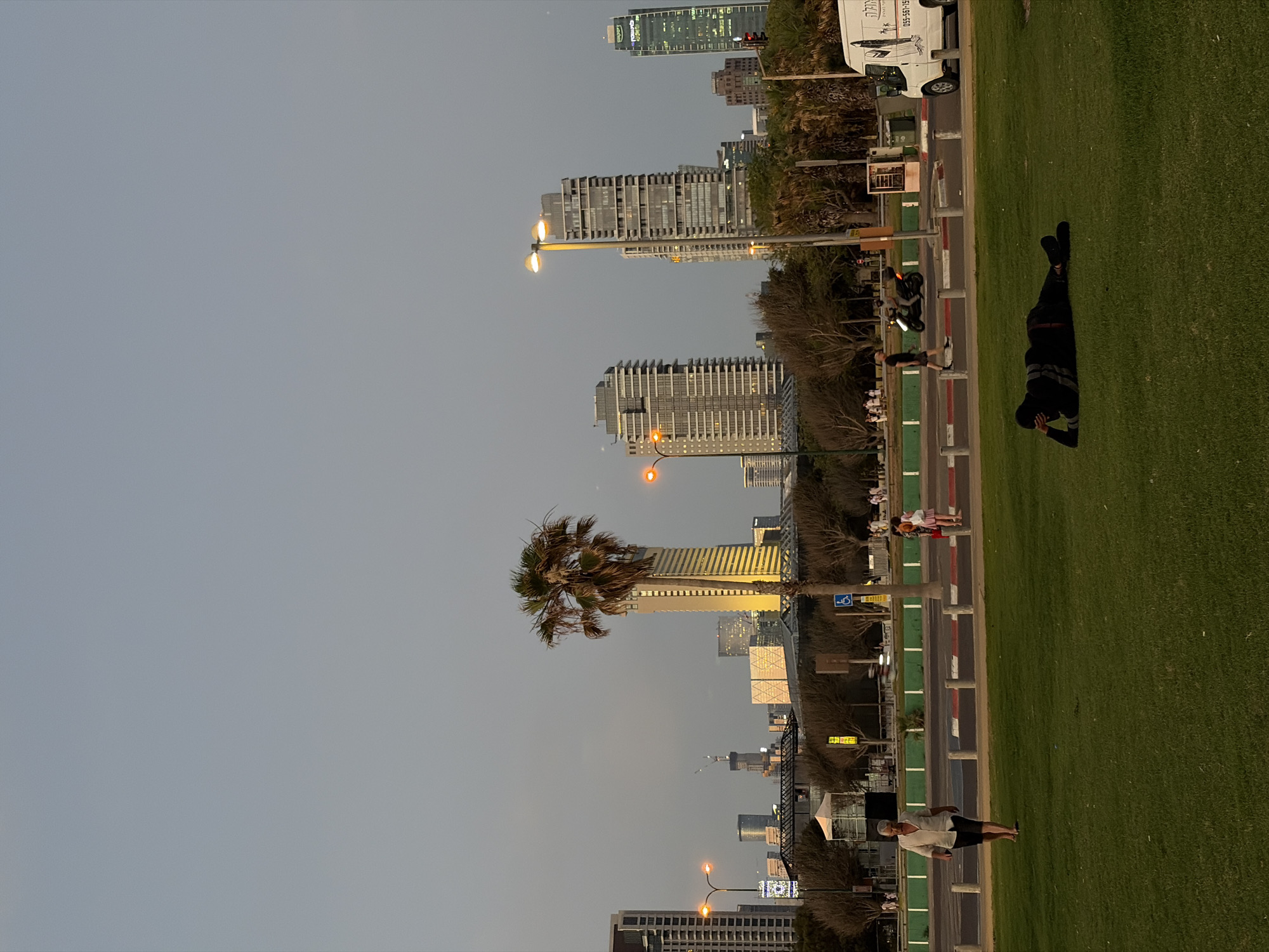 The image depicts a cityscape in Tel Aviv, Israel, likely in the late afternoon or early evening, given the soft lighting and clear sky. The foreground features a well-maintained green lawn, where a person is lying down on their side, propped up on their elbow, with their face obscured by their arm. They are wearing dark clothing, including a long-sleeved top and pants, with black shoes.
In the middle ground, a paved walkway runs parallel to the lawn, bordered by a green striped barrier and bollards. Several people are walking along the walkway. To the left, a man in a light-colored short-sleeved shirt and dark shorts walks barefoot towards the camera. Further along the walkway, two women are walking away from the camera, one wearing a pink skirt and the other a dark top and possibly shorts. A motorcycle is seen passing by in the background. A white van with graphics on its side is parked on the right side of the walkway.
The background is dominated by the Tel Aviv skyline, featuring numerous modern high-rise buildings of varying heights and architectural styles. Notable structures include a tall, slender building with a palm tree in front of it, illuminated by golden light. Other buildings are visible in the distance, some with lit windows. Streetlights are also present, with a distinct orange glow. Trees with sparse foliage line the area between the walkway and the buildings. A small white tent is visible amongst the trees. The overall environment suggests an urban park or promenade area adjacent to a business or residential district.