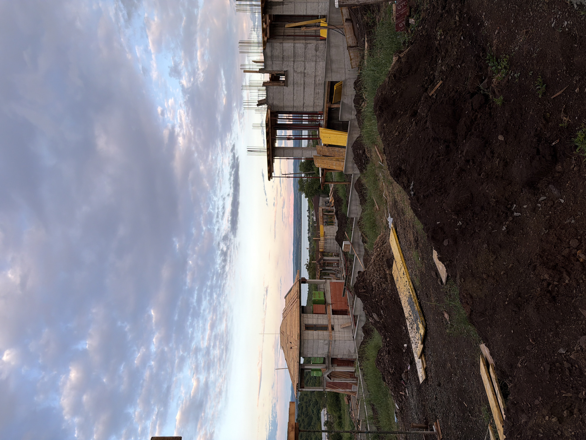 The image shows a construction site in Cherni Vrah, Bulgaria. Multiple buildings in various stages of construction are present, constructed primarily of concrete. Rebar is visible extending from the unfinished concrete structures. The surrounding landscape includes dirt, grass, and construction debris like wooden planks. In the background, there is a glimpse of a body of water and greenery. The sky above is filled with clouds.