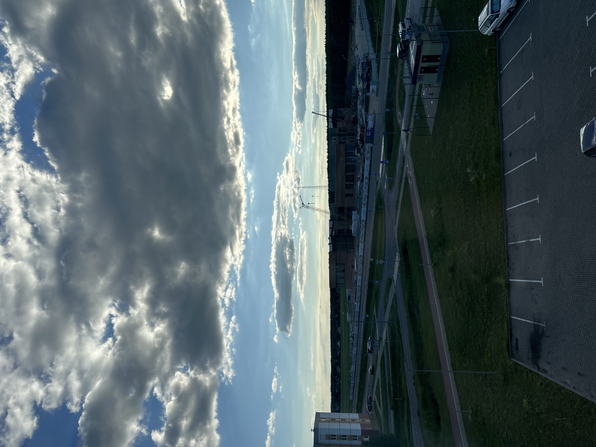 This is a vertical, eye-level, outdoor, daytime photograph taken in Pyetrykaw, Belarus. The image depicts a scene with a clear sky dominating the left side, filled with a mix of dark and light clouds. The sun is visible, casting bright light on the clouds. The right side of the image transitions to a cityscape with a parking lot, roads, green spaces, and buildings under construction.
The construction area features several cranes and buildings in various stages of completion, suggesting an ongoing development project. A paved parking lot with marked spaces is visible on the far right, with a few cars parked. Adjacent to the parking lot is a strip of grass. Roads and pathways crisscross the scene, with some traffic and pedestrian activity suggested by the presence of cars and road signs.
A prominent feature is a two-story building with a light-colored facade and visible windows. The sky's bright illumination and the presence of clouds suggest fair weather, likely late afternoon or early evening given the long shadows and the angle of light. The overall mood is one of a developing urban area against a dramatic sky. No discernible text is visible in the image.