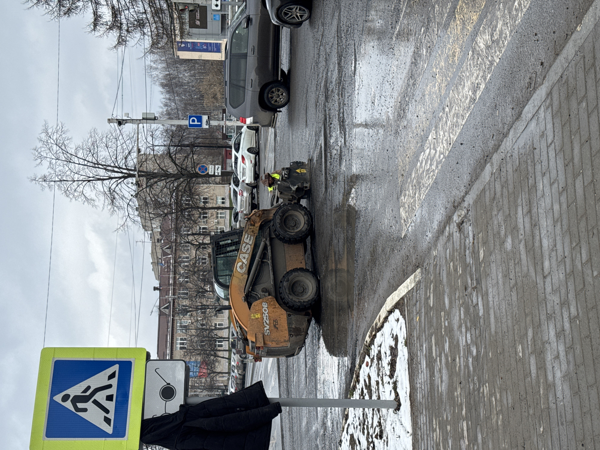 The image shows a street scene in what appears to be the city of Chaikovsky, Russia. On the left, there's a blue and yellow pedestrian crossing sign and a smaller sign with a symbol suggesting a hearing aid, possibly indicating accessibility. A CASE SV250B skid-steer loader is parked on the road. A person wearing a yellow vest stands near a smaller machine behind the loader. Several parked cars are visible along the street. The environment looks wet, with puddles on the road and patches of snow along the sidewalk, indicative of recent or ongoing snowmelt. Buildings are visible in the background.