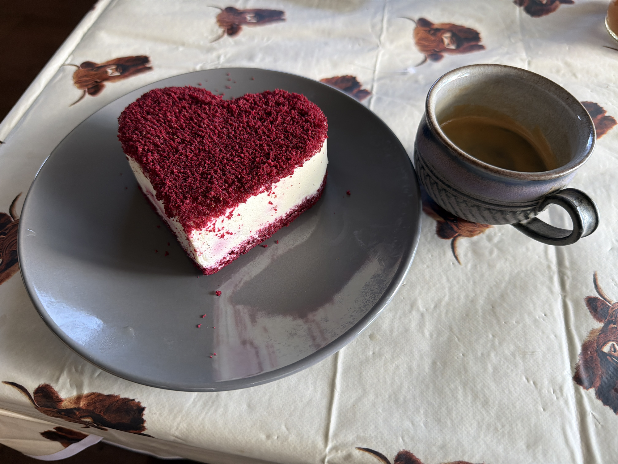 A heart-shaped slice of red velvet cake with white frosting sits on a gray plate. The cake is partially covered in red cake crumbs. A dark-colored mug, with a rough texture and a handle, is placed to the right of the plate. The mug appears to be partially filled with a dark liquid. Both the plate and the mug rest on a tablecloth that features a repeating pattern of Highland cattle illustrations. The overall lighting suggests an indoor setting, possibly a kitchen or dining area. The image does not contain any discernible landmarks or features that would allow for the precise determination of its location within Malvern Hills, United Kingdom.