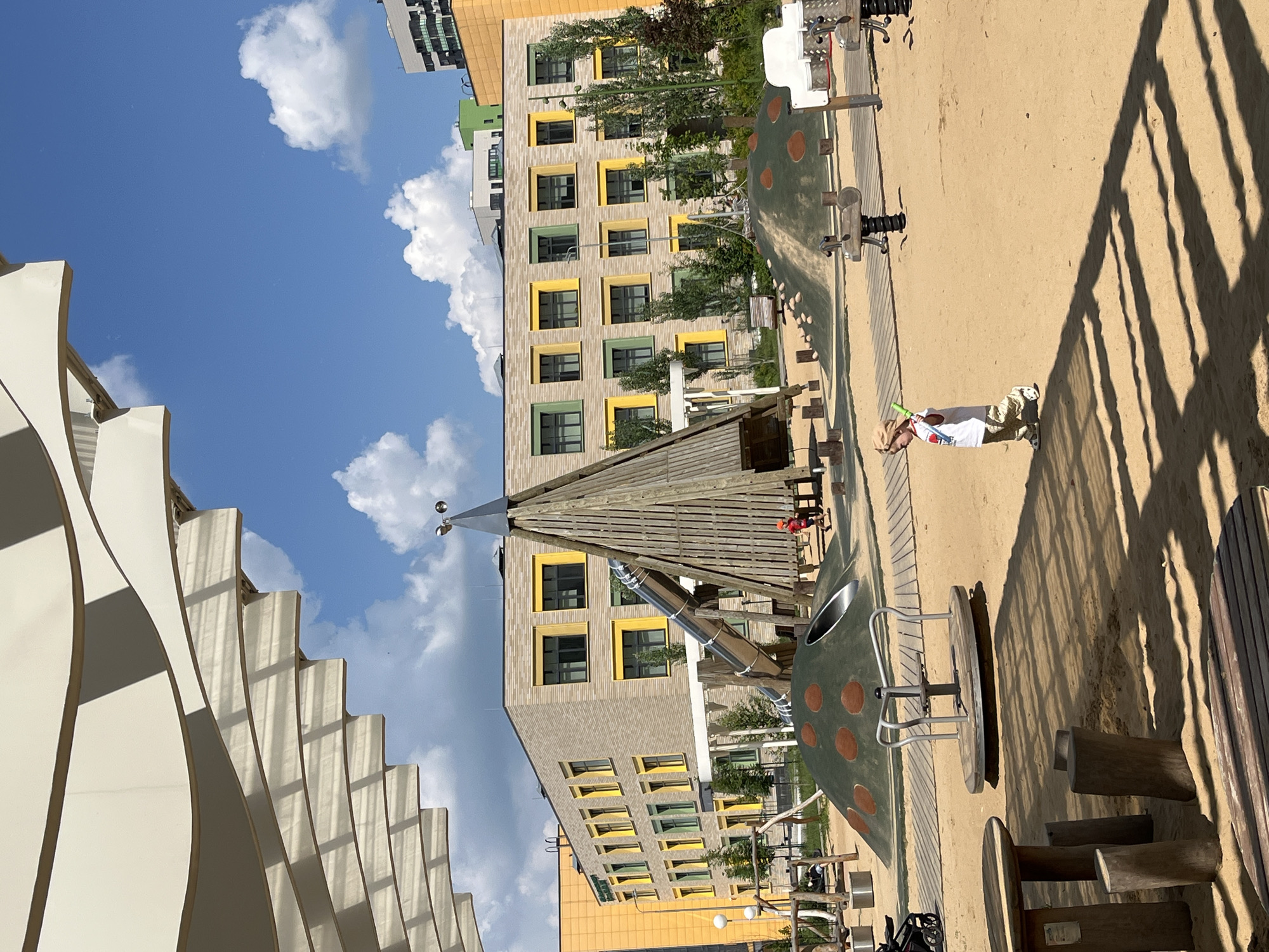 A child in a tan hat, white t-shirt, and patterned pants walks to the right across a sandy playground. The child holds a green toy in their right hand. In the background, a large, modern building with yellow and green window frames stands under a blue sky with white clouds. A tall, conical wooden structure with a metal slide is located in the foreground. The playground features a merry-go-round, wooden tables and benches, and a green, dome-shaped play structure with brown spots. The image captures an outdoor scene in a city.