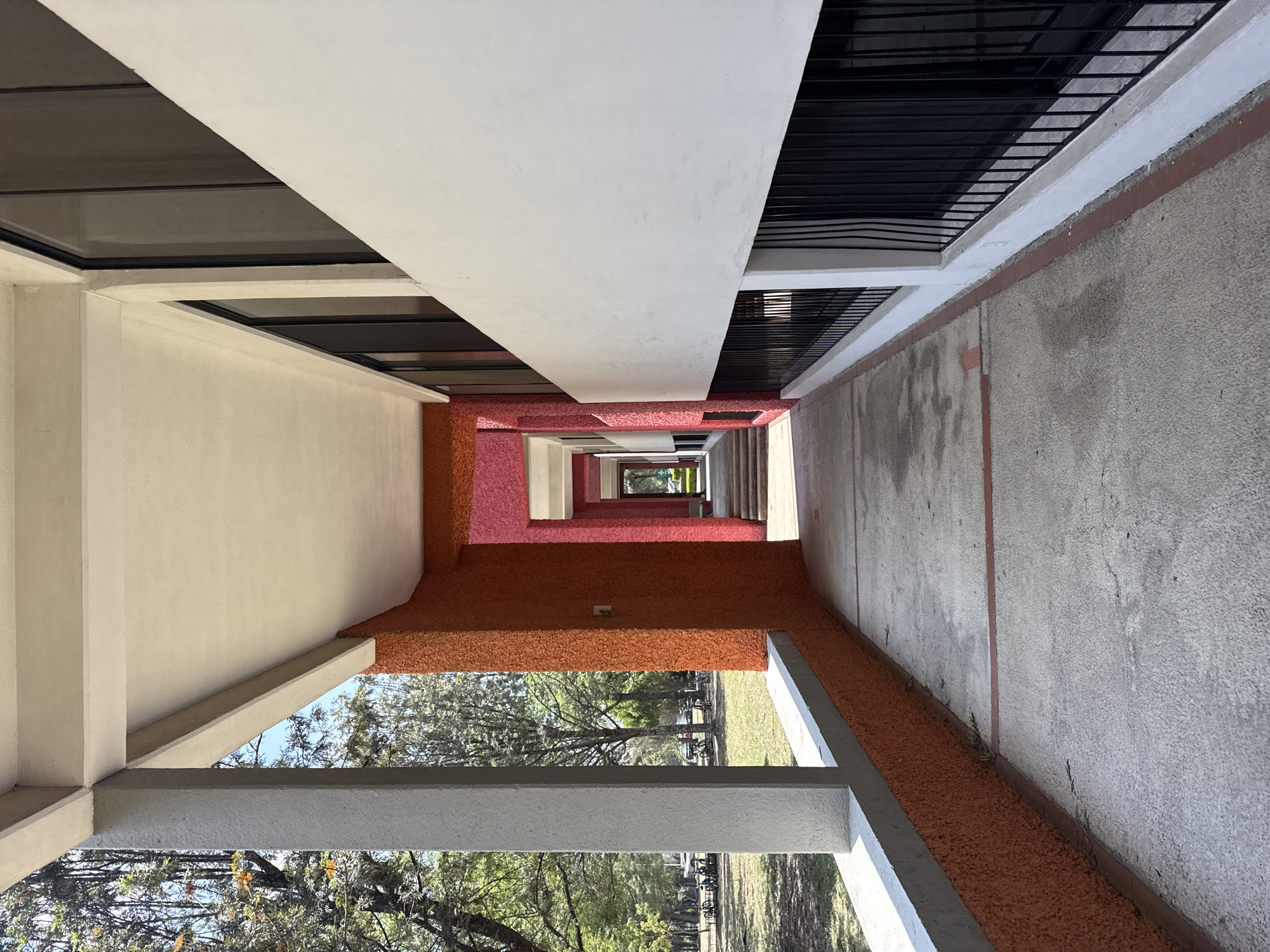 Here is a detailed description of the image:
The image captures a long, narrow perspective of a covered walkway or corridor, characterized by alternating sections of textured orange and smooth white walls and ceilings. On the left side, the structure is lined with windows, while the right features a concrete wall with occasional railings. The walkway appears to extend into the distance, terminating in a glimpse of greenery, suggesting an outdoor space beyond. The architectural style indicates the location is probably in San Andrés Cholula, Mexico.