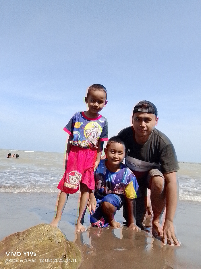 This image depicts three individuals on a sandy beach under a clear blue sky, presumably in Limbangan, Indonesia. In the foreground, from left to right, an older child stands barefoot, wearing a blue t-shirt with a space-themed graphic and red shorts with a white emblem, looking at the camera. Centered in front of an adult male, a younger child is crouched barefoot, wearing a blue and yellow t-shirt with a multi-character graphic and blue shorts, also looking at the camera. To the right, an adult male is in a crouched position, barefoot, wearing a dark t-shirt, dark shorts, and a backward-facing black baseball cap, looking at the camera with a faint smile. His left hand rests on the wet, reflective sand. In the lower-left foreground, a dark, irregularly shaped object (possibly a rock or driftwood) sits on the wet sand. The background features a calm sea with visible waves and two indistinct individuals partially submerged in the water further out to the left. The sky is bright and cloudless. The watermark "vivo Y19s Akhtar & Arvan 12 Okt 2025, 14.11" is visible in the lower-left corner.