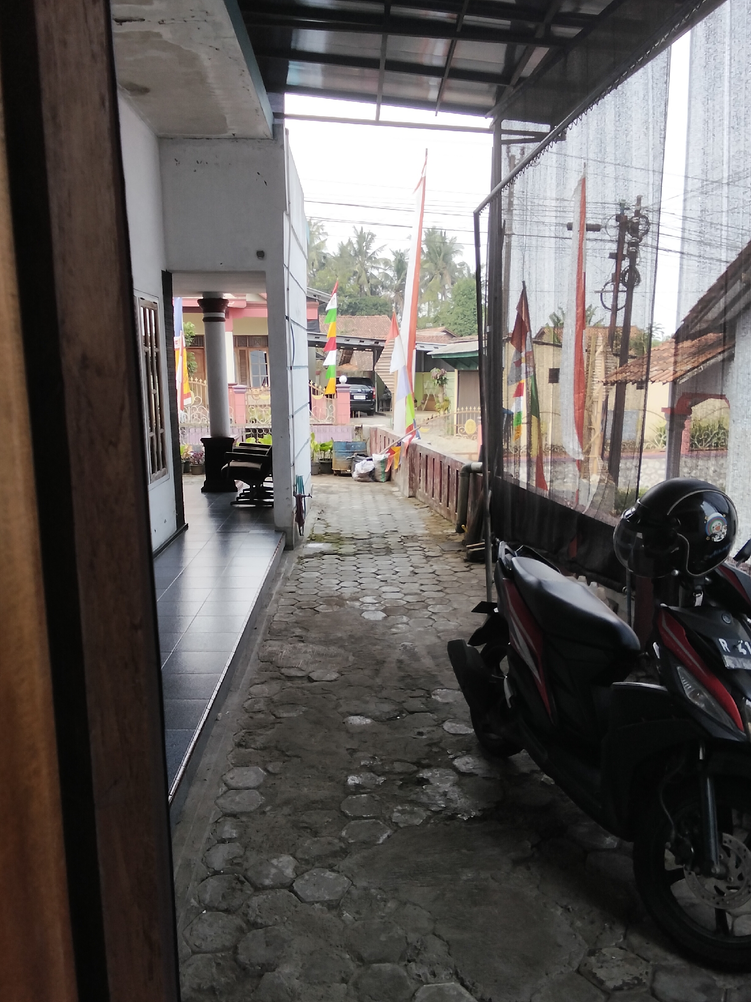 The image depicts a view of a pathway alongside a building in Paguyangan, Indonesia. The pathway is paved with hexagonal concrete blocks and is lined with a low red wall on the right. A black scooter with a red stripe and a black helmet on its seat is parked on the pathway. Behind the wall, a black car is visible on a street. Several Indonesian flags are displayed along the pathway, some attached to poles and others hanging from a fence. The building on the left has a tiled floor walkway and a white exterior wall. The overall scene appears to be an outdoor area of a residential or commercial property.