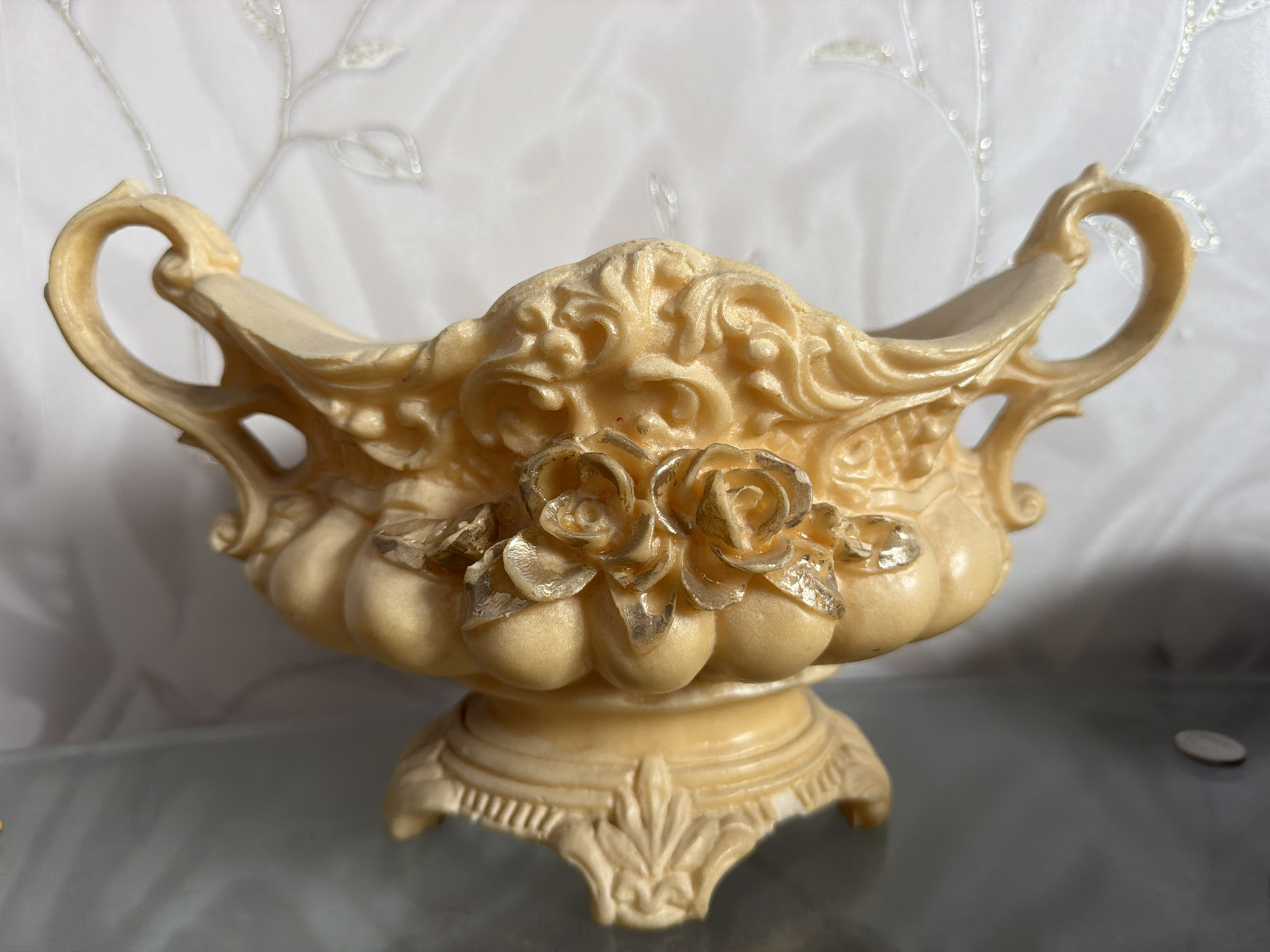 An ornate, cream-colored, decorative bowl with two handles and a pedestal base is displayed on a reflective surface. The bowl features intricate floral carvings and is adorned with metallic gold roses. The background consists of a white, slightly translucent fabric with a subtle, embroidered or printed vine pattern. A small, silver-colored coin rests on the reflective surface to the right of the bowl. The overall scene is lit from the front and slightly from above, creating soft shadows. The image was captured in Yuzhno-Sakhalinsk, Russia.