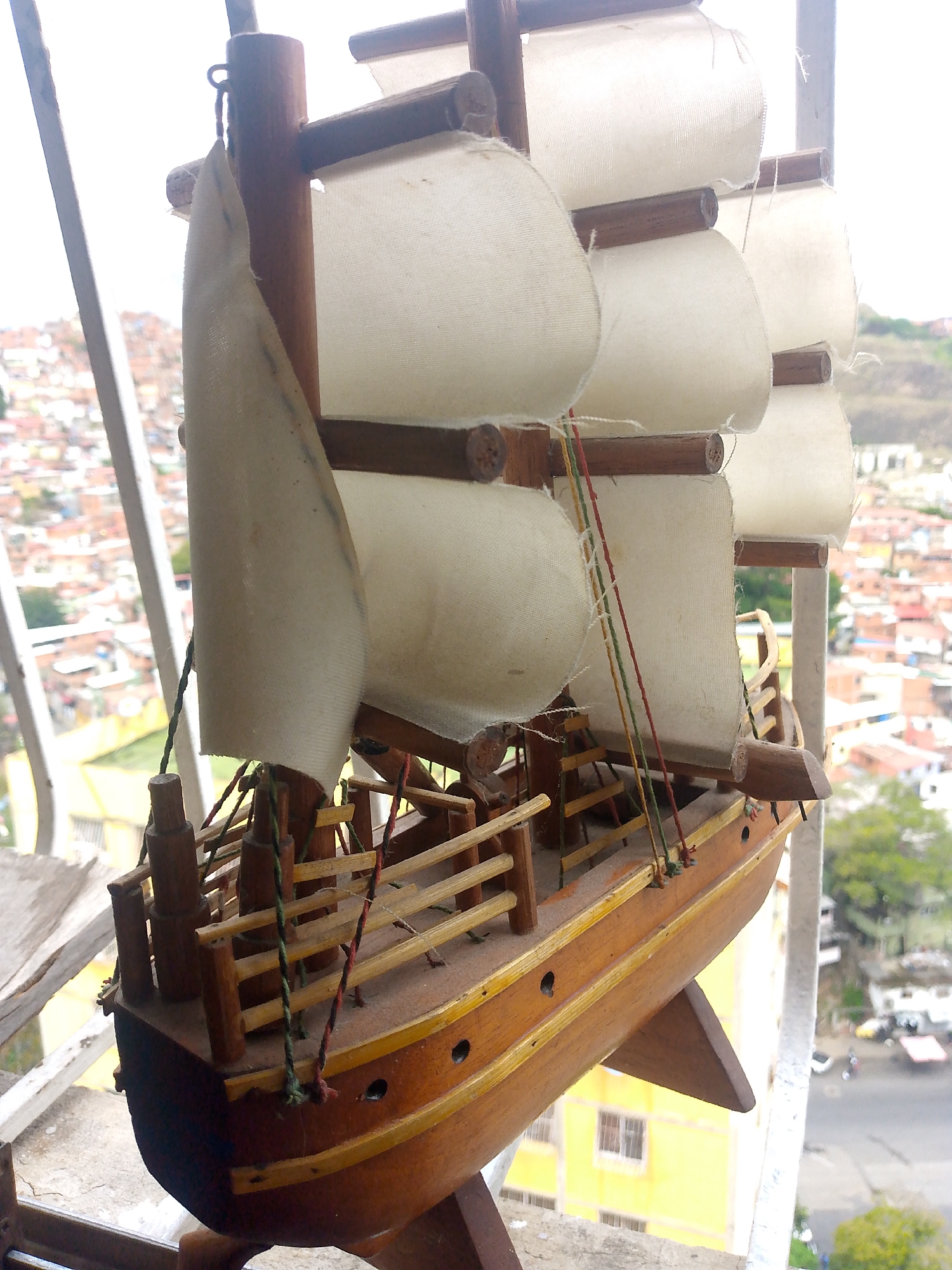 The image shows a model wooden ship with white cloth sails, displayed on a balcony. The ship has brown wooden masts, a brown hull with small round windows, and a wooden railing around the deck. Red, green, and yellow string adorn the ship. In the background, a cityscape can be seen, with densely packed houses and buildings in the distance. The location is Caracas, Venezuela, as evidenced by the characteristic urban sprawl and architectural style visible in the cityscape. The image captures a blend of the handmade model ship with a backdrop of the city.