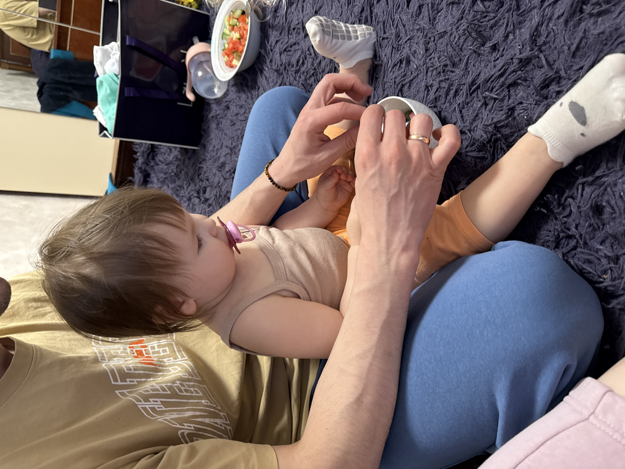 A baby is lying on a person's lap, who is sitting on a dark gray carpet. The baby has brown hair and is wearing a beige tank top and orange shorts. The baby has a pink pacifier in their mouth. The person holding the baby is wearing blue sweatpants and a gold ring on their finger. They have a bowl of food on the carpet in front of them and they seem to be feeding the baby. A bag and a bottle are sitting next to the bowl. The scene is located in an interior space in Krapivka, Russia.