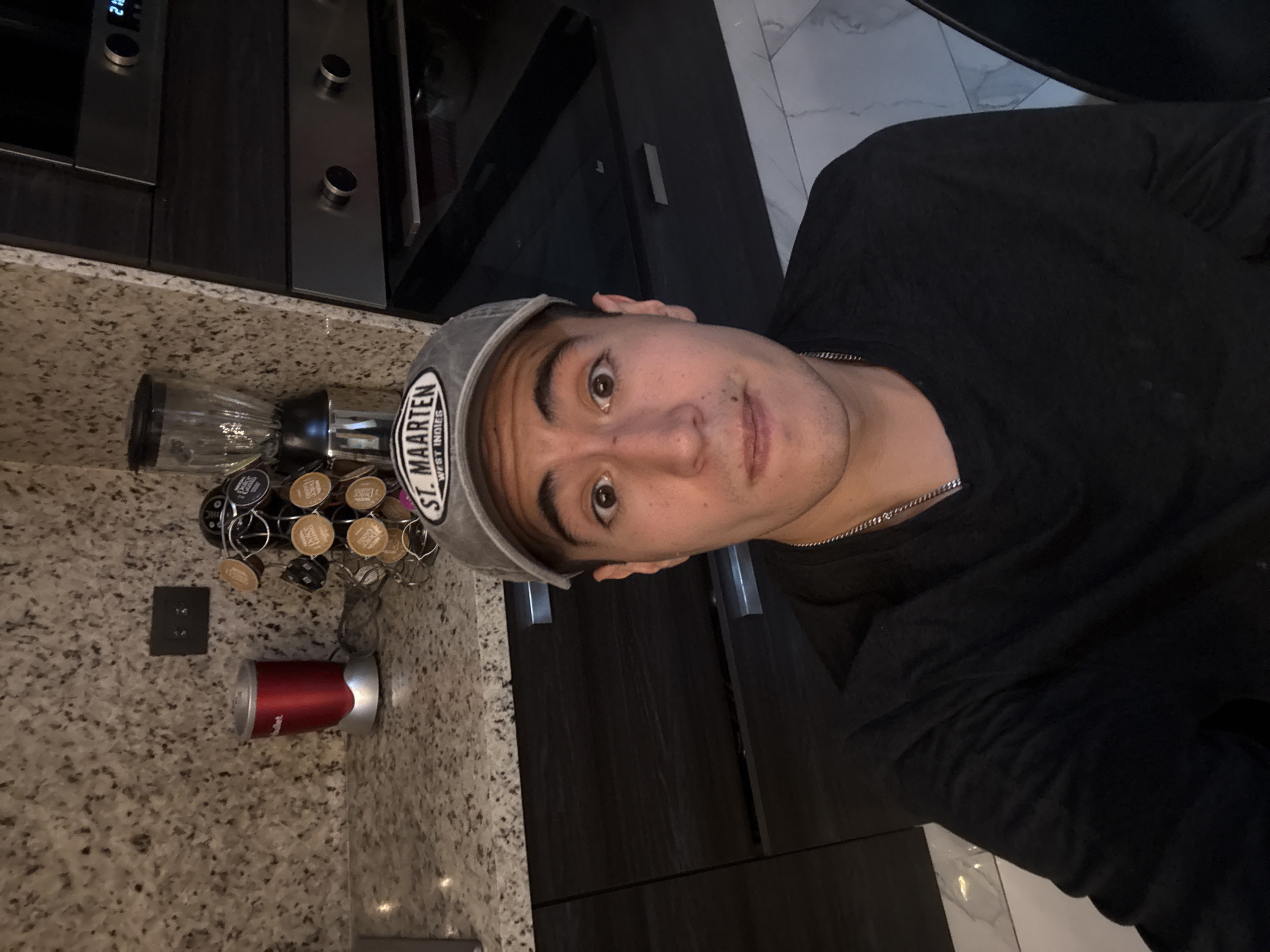 A person wearing a grey baseball cap with "ST. MAARTEN" printed on it and a black long-sleeved shirt is in a kitchen. The kitchen has granite countertops, dark cabinets, and stainless steel appliances. There's a coffee pod carousel with various coffee pods and a red and silver coffee machine on the counter. The floor is tiled in a marble pattern. The specific location within San Bernardino Tlaxcalancingo, Mexico, is not discernible from the image.