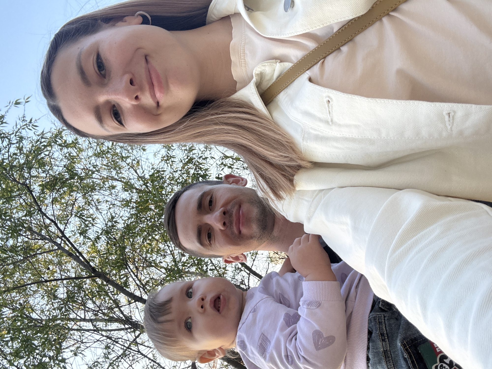 A selfie shows a woman, a man, and a baby in an outdoor setting. The woman is at the top of the frame, smiling. The man is below her, also smiling. The baby is at the bottom, looking at the camera. The background includes trees and a blue sky. The location is not discernible as Moscow without additional visual cues or context.