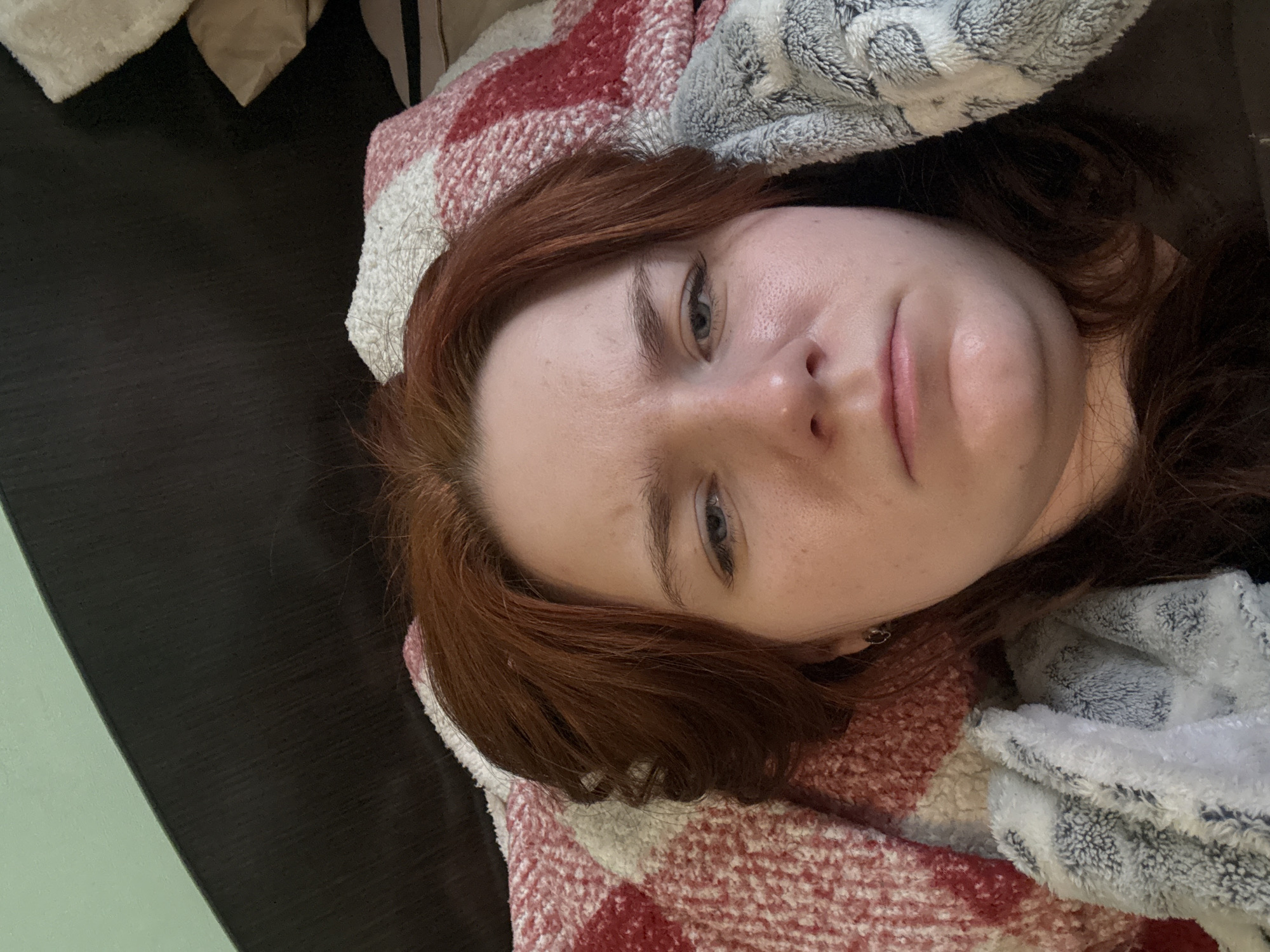 A close-up, low-angle shot depicts a person lying down on what appears to be a bed. The individual has fair skin, reddish-brown hair, and light-colored eyes. They are partially covered by a blanket or quilt with a red and white checkered pattern, as well as a soft, gray and white blanket. The background features a dark-colored surface, possibly the bed frame, and a glimpse of a light green or blue wall. The city and country are not discernible from the image.