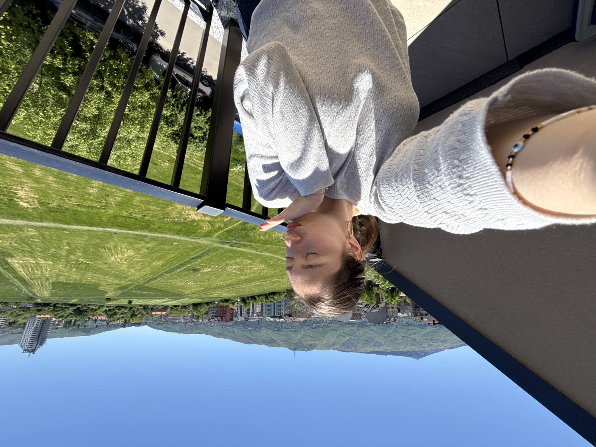 A medium shot shows a person on a balcony, with the city of Sofia, Bulgaria in the background. The person is wearing a light gray sweater and has a beaded bracelet on their wrist. They are holding their hand near their mouth. The background includes a large grassy field, buildings, mountains, and a clear blue sky. A black railing is visible on the balcony.