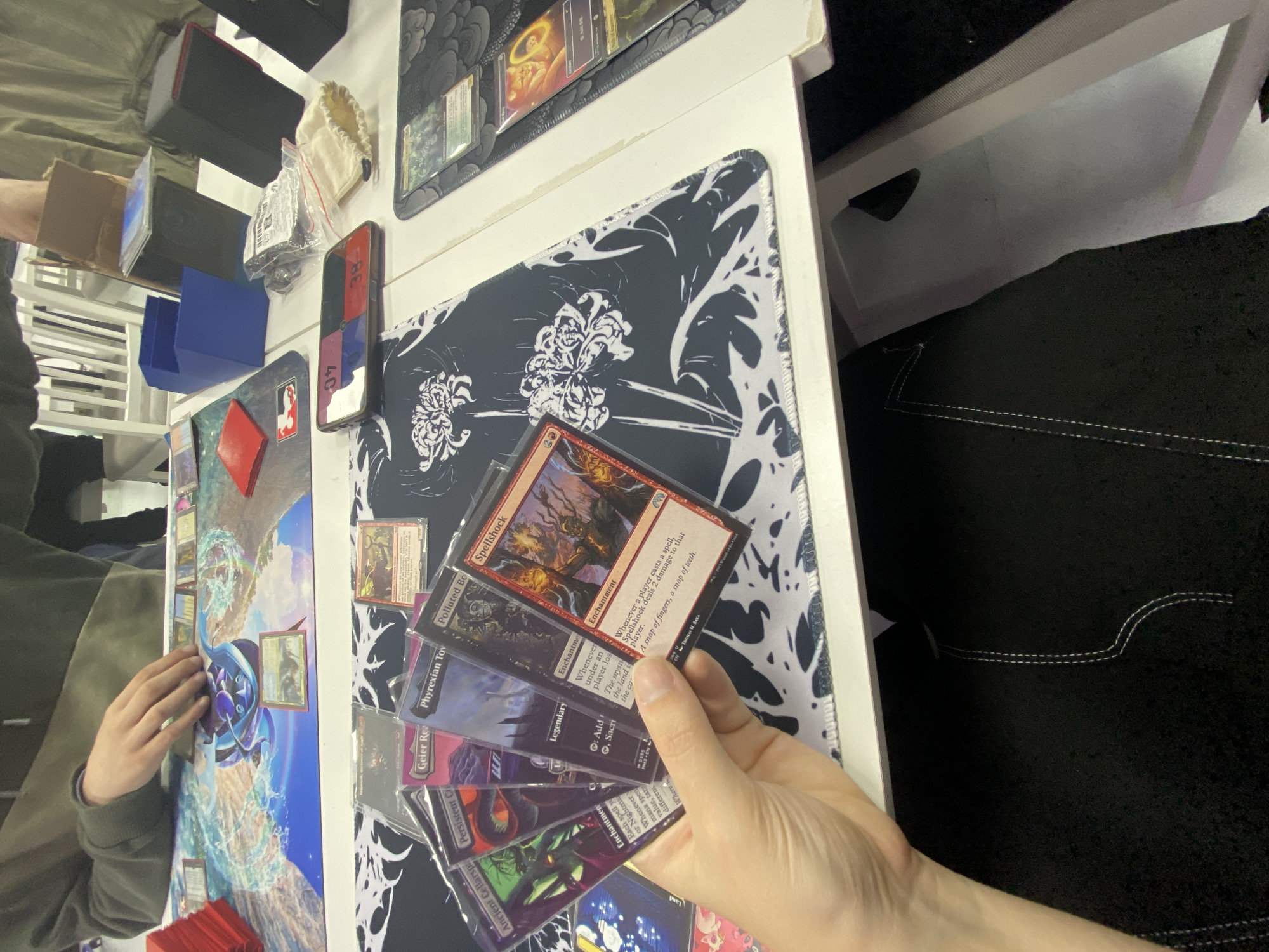 The image depicts a game of Magic: The Gathering in progress. A person is holding a hand of cards, including "Ancient Cellarspa", "Persistent Cryptolith", "Geier Reach Sanitarium", "Phyrexian Tower", "Polluted Bonds", and "Spellshock". Cards are laid out on a playmat, and the edges of the playing surface are visible. Other items are present such as card boxes, dice, and accessories. It is not possible to determine a specific location in the city of Gijón/Xixón, Spain from the image alone.