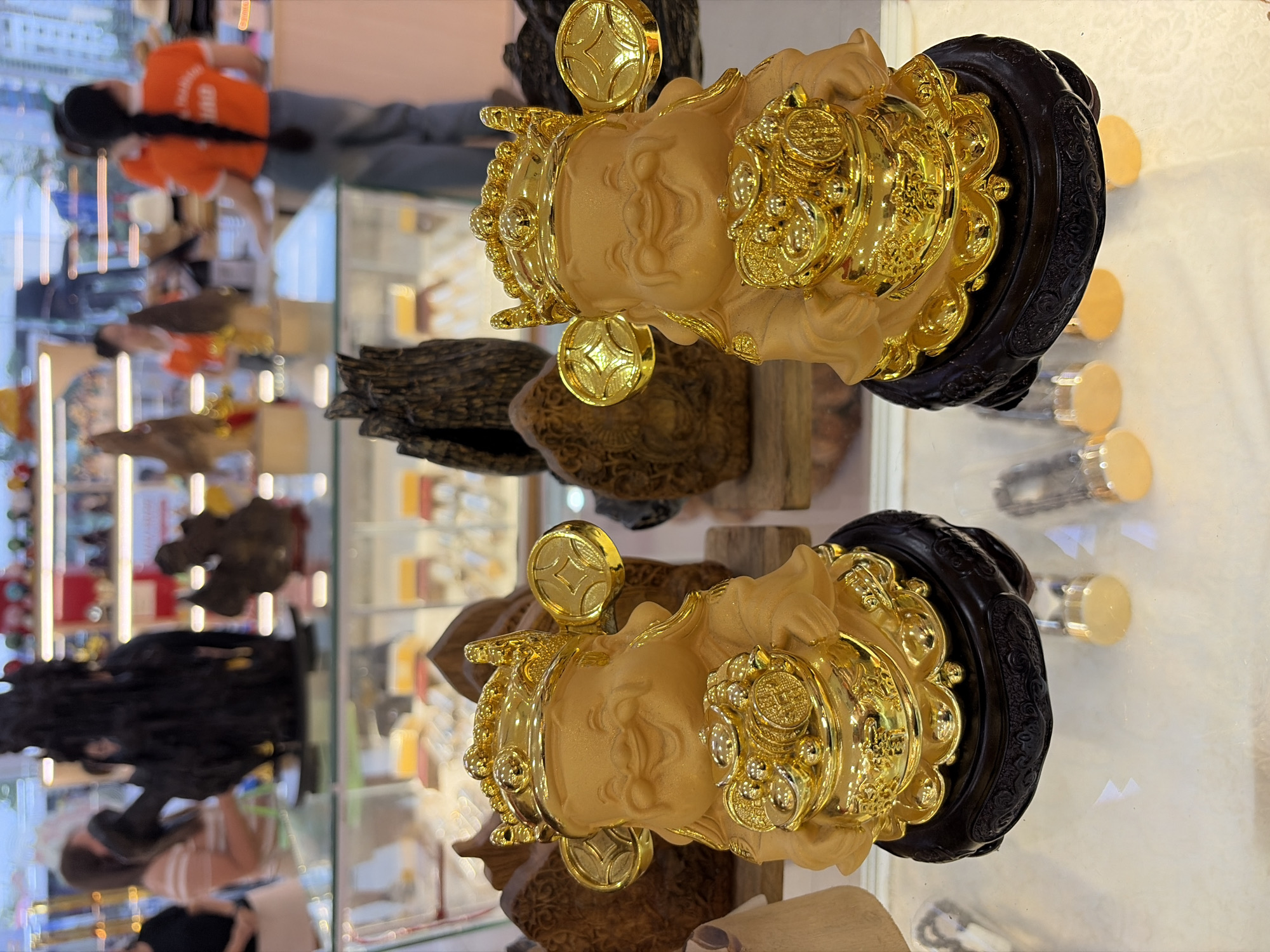 The image displays two golden figurines of a smiling, plump figure wearing a crown and holding a pot overflowing with gold coins. The figurines are positioned on dark, ornate bases, which rest on a reflective surface. Behind the figurines, various wooden decorative items and shelves stocked with merchandise are visible, suggesting a retail environment. In the background, blurred figures of people can be seen, some wearing bright orange shirts, indicating they are likely employees or customers in a store. The overall scene suggests a display of decorative or auspicious items within a shop. Based on the context provided about Đà Nẵng, Vietnam, this scene could be found in a souvenir shop or a market specializing in decorative goods in the city.