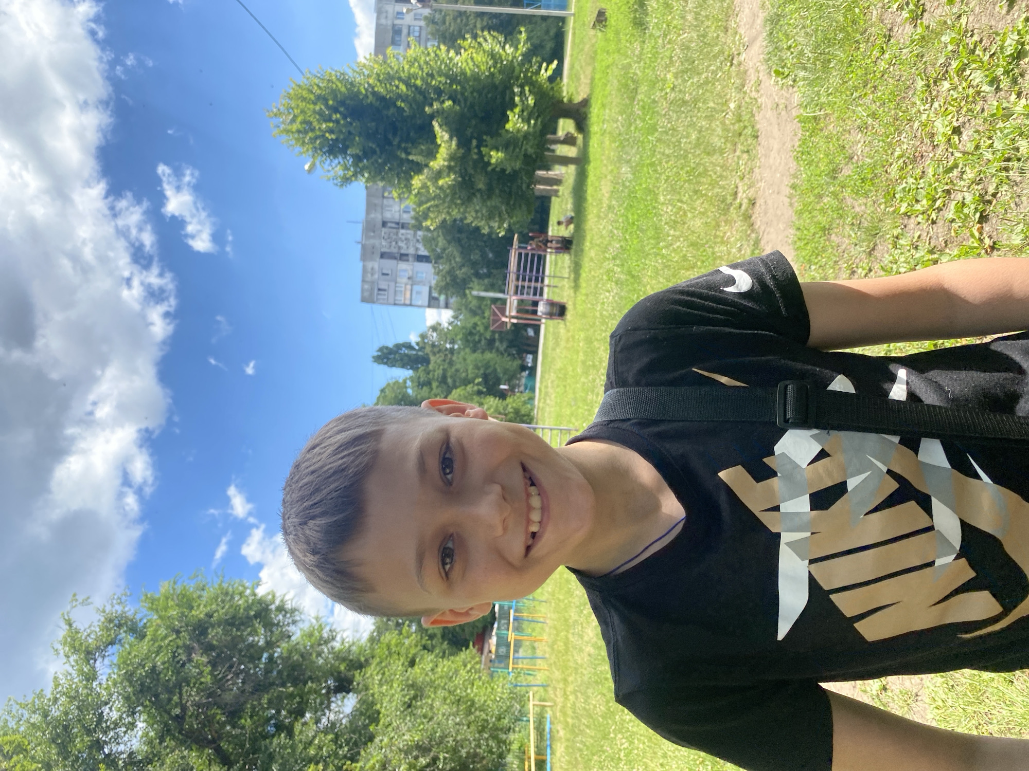 The media file shows a young boy smiling at the camera in an outdoor setting. He is wearing a black t-shirt with a graphic design that appears to be the "Nike" logo. The boy has short, dark hair and is looking directly at the viewer with a wide, toothy grin.
The background suggests a park or playground area, with lush green grass, trees, and what looks like part of a playground structure. In the distance, a multi-story building is visible, characteristic of an urban environment. The sky is blue with scattered white clouds, indicating fair weather and likely daytime. The orientation of the photo is slightly tilted.
There is visible text on the boy's t-shirt, appearing to be the Nike logo in gold, white, and grey. The overall mood is cheerful and casual, captured on a bright day. The location context is provided as Kharkiv, Ukraine.