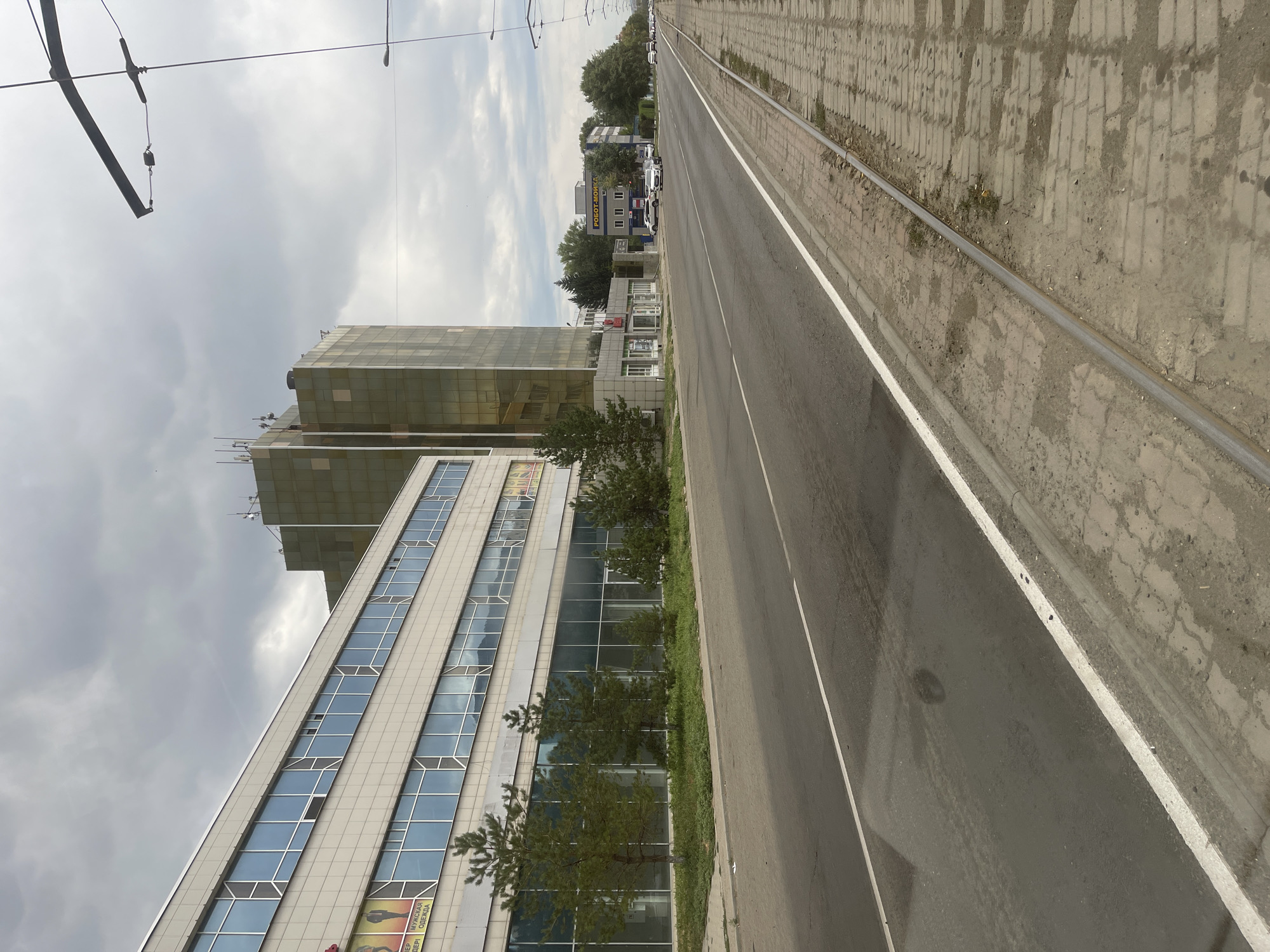 The image captures a view of a street in the city of Oskemen, Kazakhstan. The street is asphalted and has a tram track running along the right side, bordered by a paved sidewalk. On the left side of the street, there is a modern building with a facade of light-colored panels and large glass windows, some of which are open. In front of this building, several evergreen trees are planted. Further down the street, to the right of the first building, stands a taller, modern building with a distinctive golden-tinted glass facade. Beyond this, more buildings and trees line the street, with several vehicles visible on the road. The sky is overcast with clouds. The overall scene depicts a typical urban environment on a cloudy day.