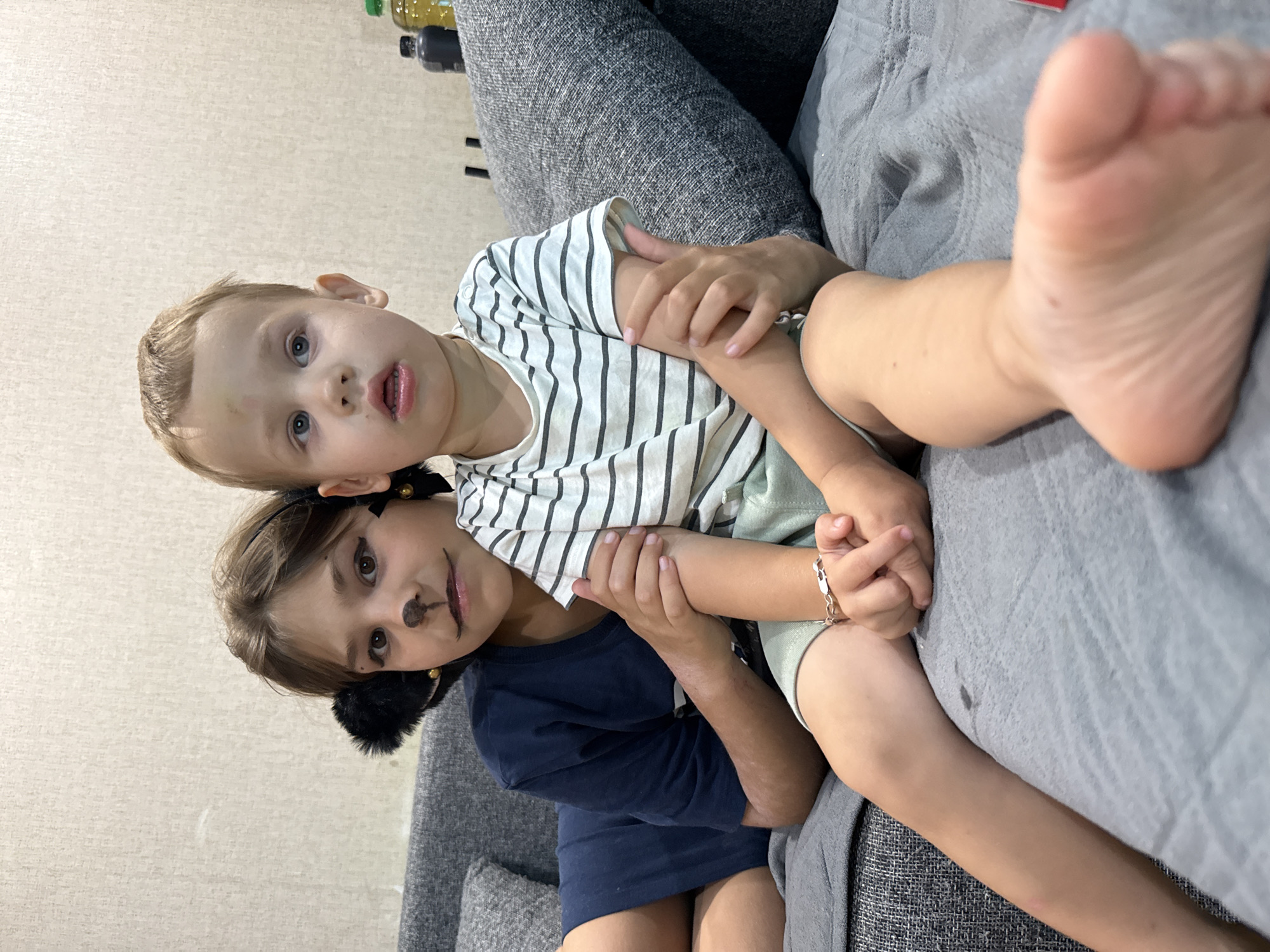 Two young children are sitting on a grey couch in what appears to be a domestic setting. The child on the left, possibly a girl due to the makeup resembling a cat's face (whiskers, nose, and eyeliner) and furry ears attached to a headband, is wearing a dark blue t-shirt. She is holding the arm of the child on the right. The child on the right, possibly a boy, is wearing a striped t-shirt and light green shorts. He is looking forward with an open mouth, and a faint bruise is visible on his forehead. Both children are resting on a grey blanket that covers the couch. The background consists of a textured, light-colored wall. Bottles of liquid are visible on a surface behind the children, suggesting a living area or a room where such items might be stored. The overall scene depicts a moment of interaction between two siblings.