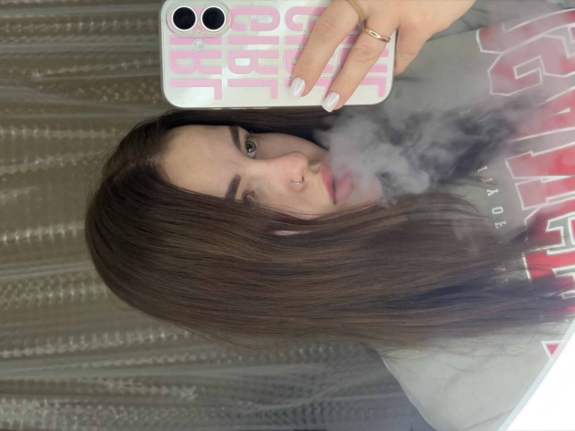 A person with brown hair is holding a white cell phone with a pink case. The case has the word "GIRL" written on it. The person has a nose ring and is exhaling smoke. The person is wearing a grey sweatshirt. The background is a light brown textured surface.