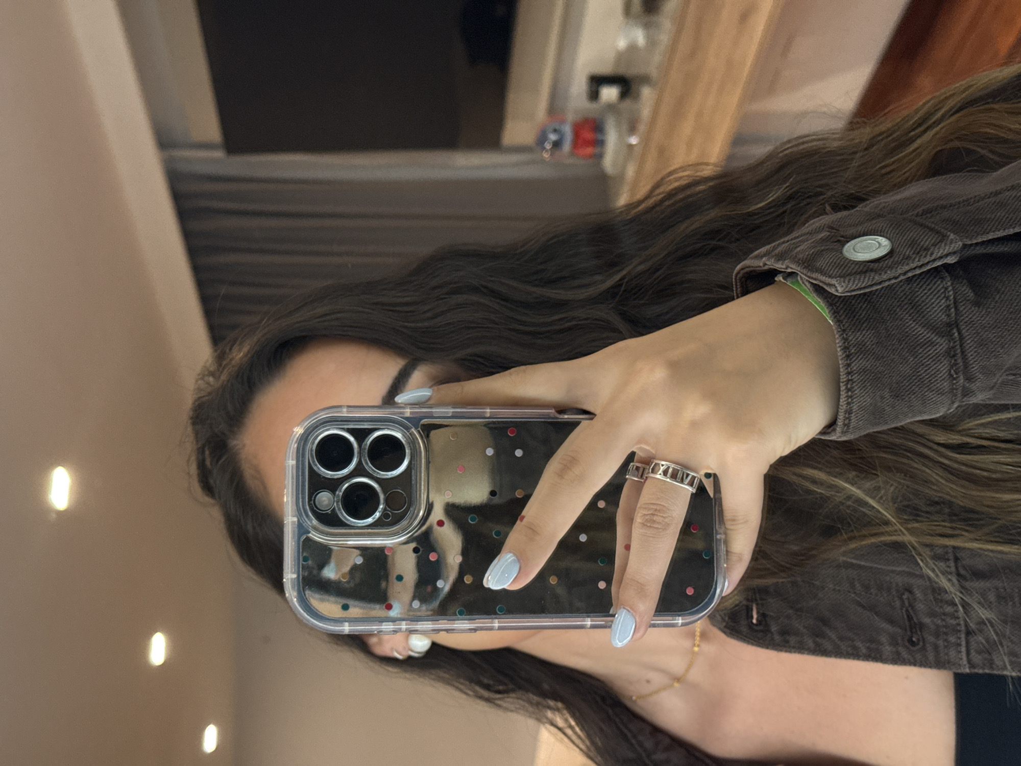 A young woman is taking a selfie in what appears to be an indoor environment, possibly a room in a home or apartment. She holds a smartphone with a clear case decorated with colorful polka dots, which obscures her face in the reflection. She has long, dark hair with some lighter strands. Her nails are painted a pale blue. She is wearing rings on one finger, and a dark brown denim jacket with a button visible on the sleeve cuff. A gold necklace is visible on her neck. The reflection in the mirror shows a glimpse of other furniture or decor in the background, including a shelf and a darker item (perhaps a television). Based on the environment being a "room in a home or apartment," it is not possible to determine the location of the city of San Bernardino Tlaxcalancingo, Mexico.