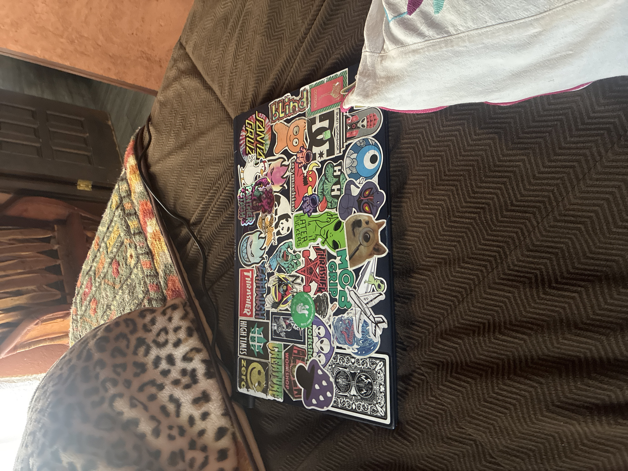 The image shows a laptop covered in stickers resting on a brown, textured surface, possibly a couch or blanket. A patterned bag, featuring leopard print and other textiles, is visible on the left side of the image. A white garment with pink trim is located on the right side. The environment is a room, potentially a living room, and a door and wooden furniture are partially visible in the background. The location is not discernible as Cañada de Madero, Mexico.
