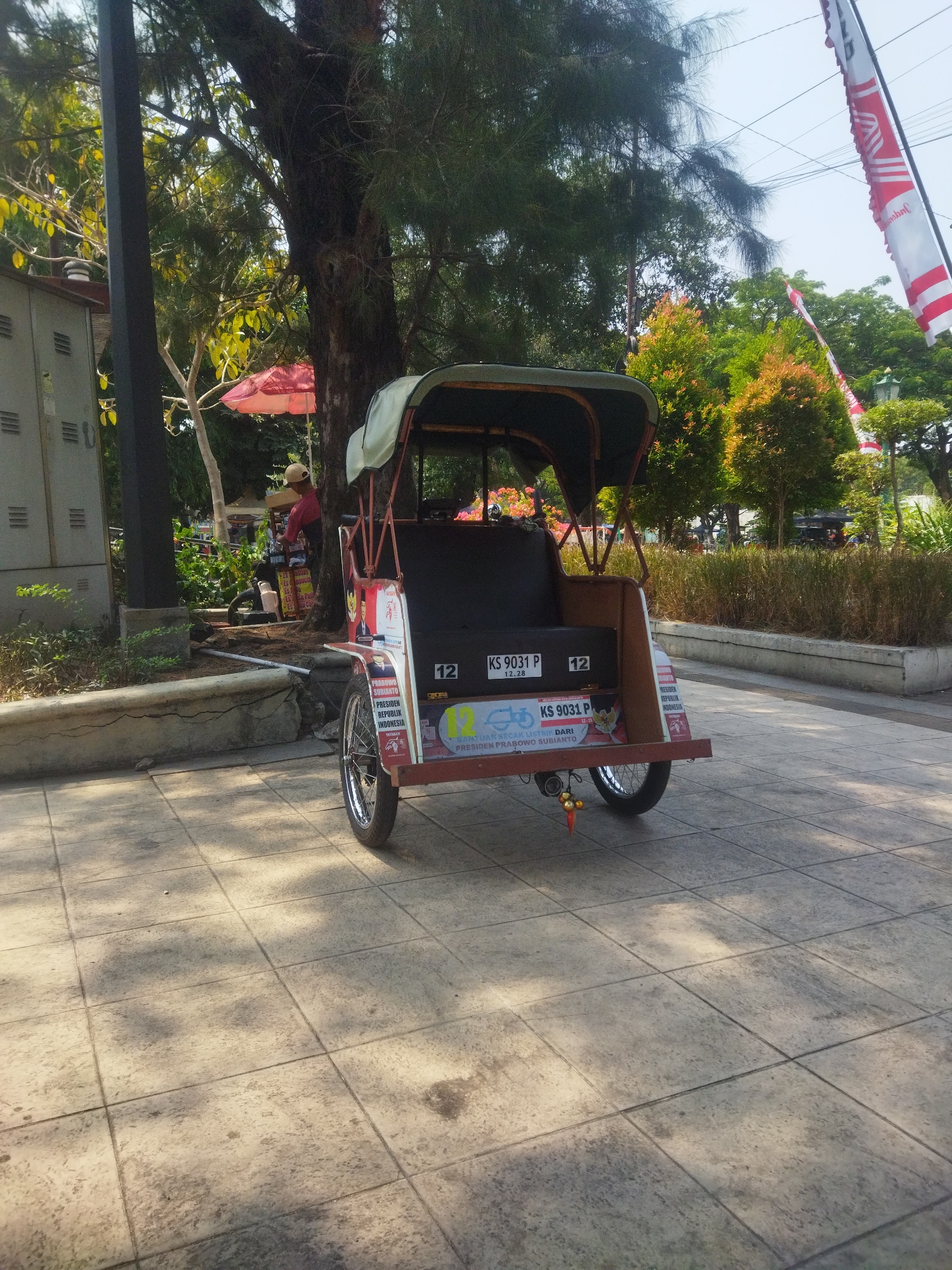 The image depicts a becak, a three-wheeled passenger cart, parked on a tiled walkway in Surakarta, Indonesia. The becak is predominantly red and black, with a green canopy. It features various stickers, including one with the text "PRABOWO SUBIANTO PRESIDEN REPUBLIK INDONESIA" and another with license plates "KS 9031 P". In the background, a person wearing a red shirt and a cap is visible near a food stall with a red umbrella. Trees and foliage surround the area, and flags, likely Indonesian flags, are seen hanging in the distance. The walkway is illuminated by sunlight, casting shadows from trees. The environment suggests a public space within the city, possibly a park or a busy street.