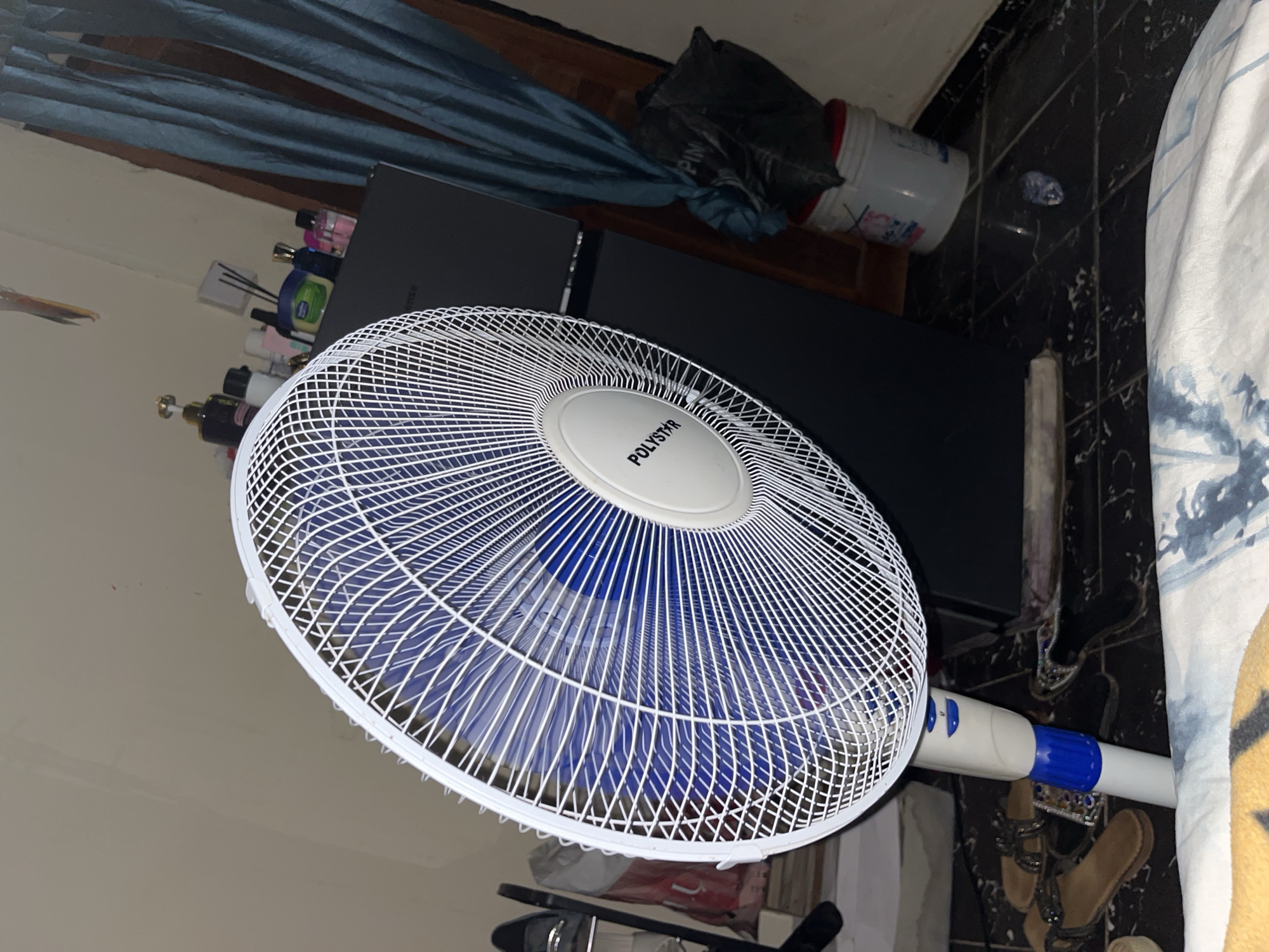 The image shows a floor fan positioned in a room, likely a bedroom or living area. The fan is white with a blue base and is actively spinning, creating a blur of its blue blades. The fan is branded "POLYSTAR". To the right of the fan, a dark grey refrigerator or cabinet is visible, topped with several bottles of what appear to be toiletries or perfumes, and a green jar. Blue curtains are drawn behind the refrigerator. On the floor in front of the fan, there are a pair of brown sandals and a dark-colored object that might be another pair of shoes or a bag. The floor is tiled with a black and white pattern. In the background, to the left, a shoe rack with shoes and a red and white bag are partially visible. A crumpled plastic bottle is on the floor in the lower right section of the image. The scene suggests a somewhat cluttered domestic environment. The presence of this type of fan and the general appearance of the room are consistent with residential settings in Jos, Nigeria, where such appliances are common for cooling.