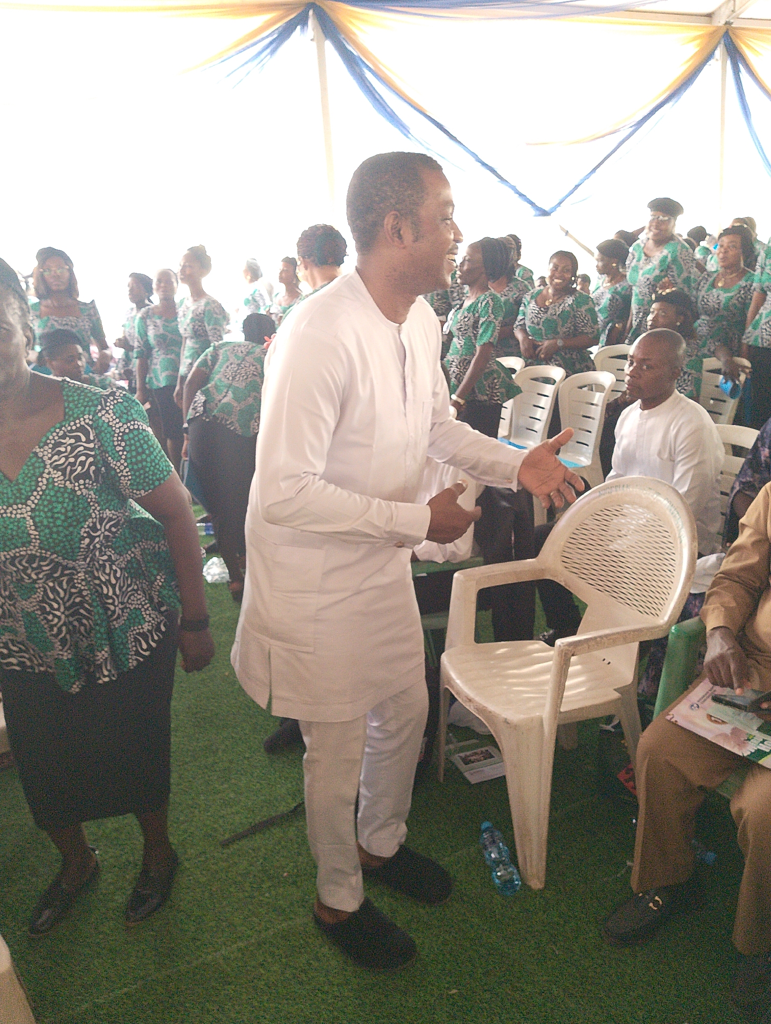The image depicts a lively gathering under a large tent, with bright natural light illuminating the scene. The ground is covered with green artificial turf. In the foreground, a man wearing a white, possibly traditional, long-sleeved tunic and trousers stands animatedly. He is smiling broadly, looking towards the left, and gesturing with both hands, appearing to be speaking or engaging with the crowd.
Numerous other individuals populate the scene. Many women are prominent, dressed in matching green, black, and white patterned tops or dresses, suggesting a uniform or group affiliation. Their expressions generally convey smiles and engagement. Some attendees are standing, while others are seated on white plastic chairs scattered throughout the area.
In the background, overhead, blue and yellow fabric drapes are visible, forming part of the tent's decoration. A plastic water bottle lies on the turf near a white chair in the lower right foreground. Another man, also in a white long-sleeved top, is seated further back, and a person in brown attire holds a booklet or papers in the bottom right corner, though any text on it is illegible. The overall atmosphere appears joyful and social, indicative of an event or meeting.