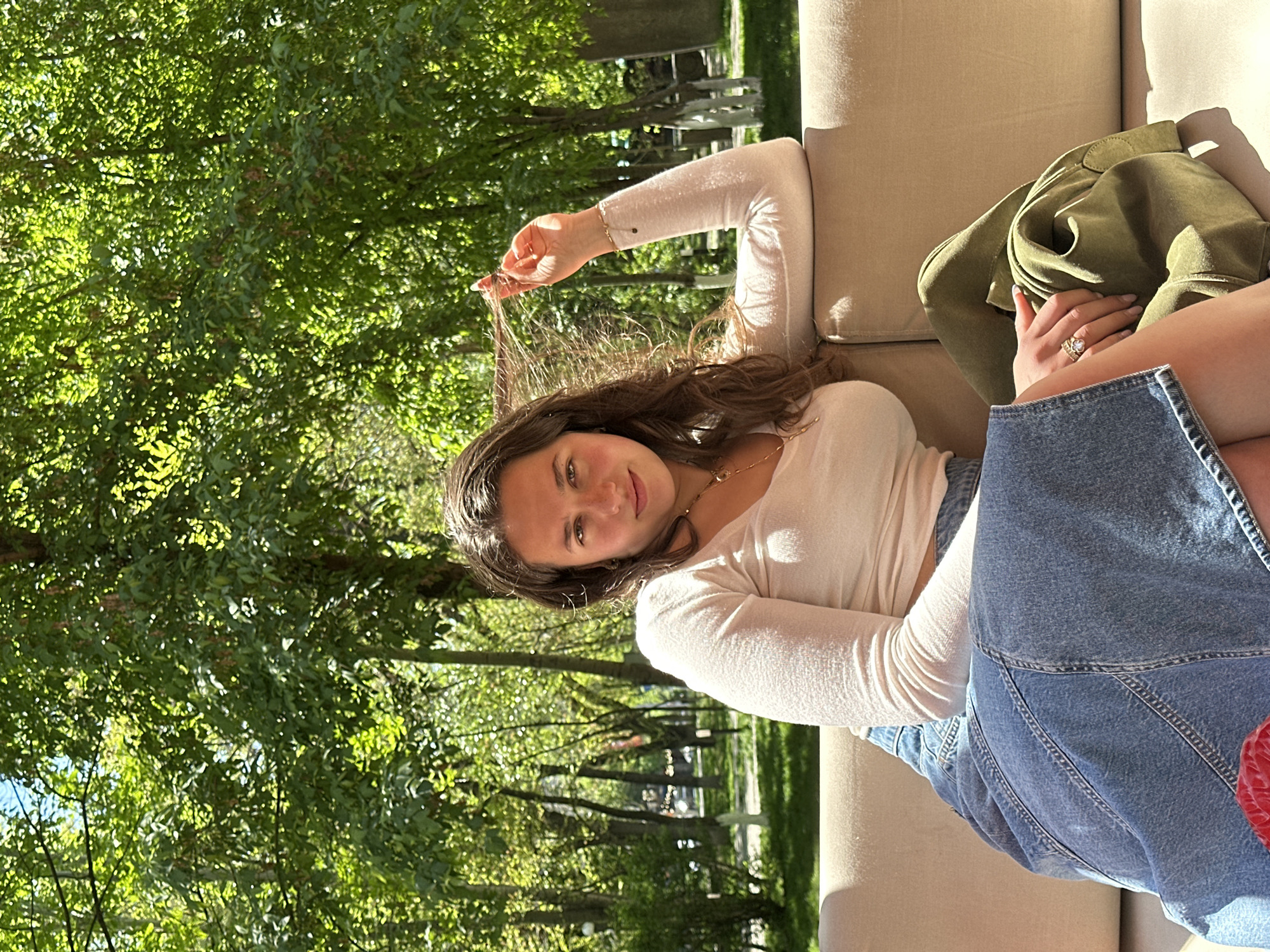 The image captures a young woman relaxing outdoors in Yerevan, Armenia. She is seen from the chest up, positioned diagonally across the frame, lying on a light-colored outdoor sofa or lounge chair. She has long, dark brown hair, a pleasant expression with a faint smile, and is looking directly at the camera. She is wearing a light cream or off-white long-sleeved top and what appears to be a denim skirt or overall bottom. A gold-toned necklace is visible around her neck, and a decorative ring is on her right hand, which is resting near her left leg. Her right arm is raised, and she is holding a strand of her hair between her fingers.
Next to her on the sofa, a green jacket or piece of clothing is casually draped. The background is dominated by lush, vibrant green foliage, indicative of numerous trees, suggesting a park, garden, or a tree-lined street. Sunlight filters through the leaves, creating bright highlights and dappled shadows, indicating a sunny day, likely morning or late afternoon given the angle of the light. The overall mood is relaxed and serene. No text is visible in the image.