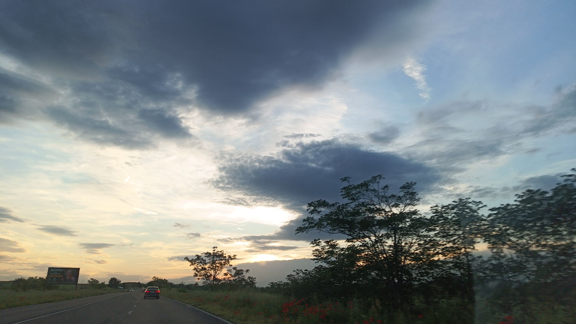 The image depicts a road scene during twilight, likely on the outskirts of Burgas, Bulgaria, although no specific landmarks confirm the city. The road stretches into the distance with a car visible ahead. The foreground is lined with trees and vegetation, including what appear to be red poppies. A billboard stands on the left side of the road. The sky is dominated by a mix of dark, heavy clouds and lighter, sunlit areas. The sun appears to be setting or rising, casting a warm glow on the horizon. No people are clearly visible, focusing on the landscape and the road.