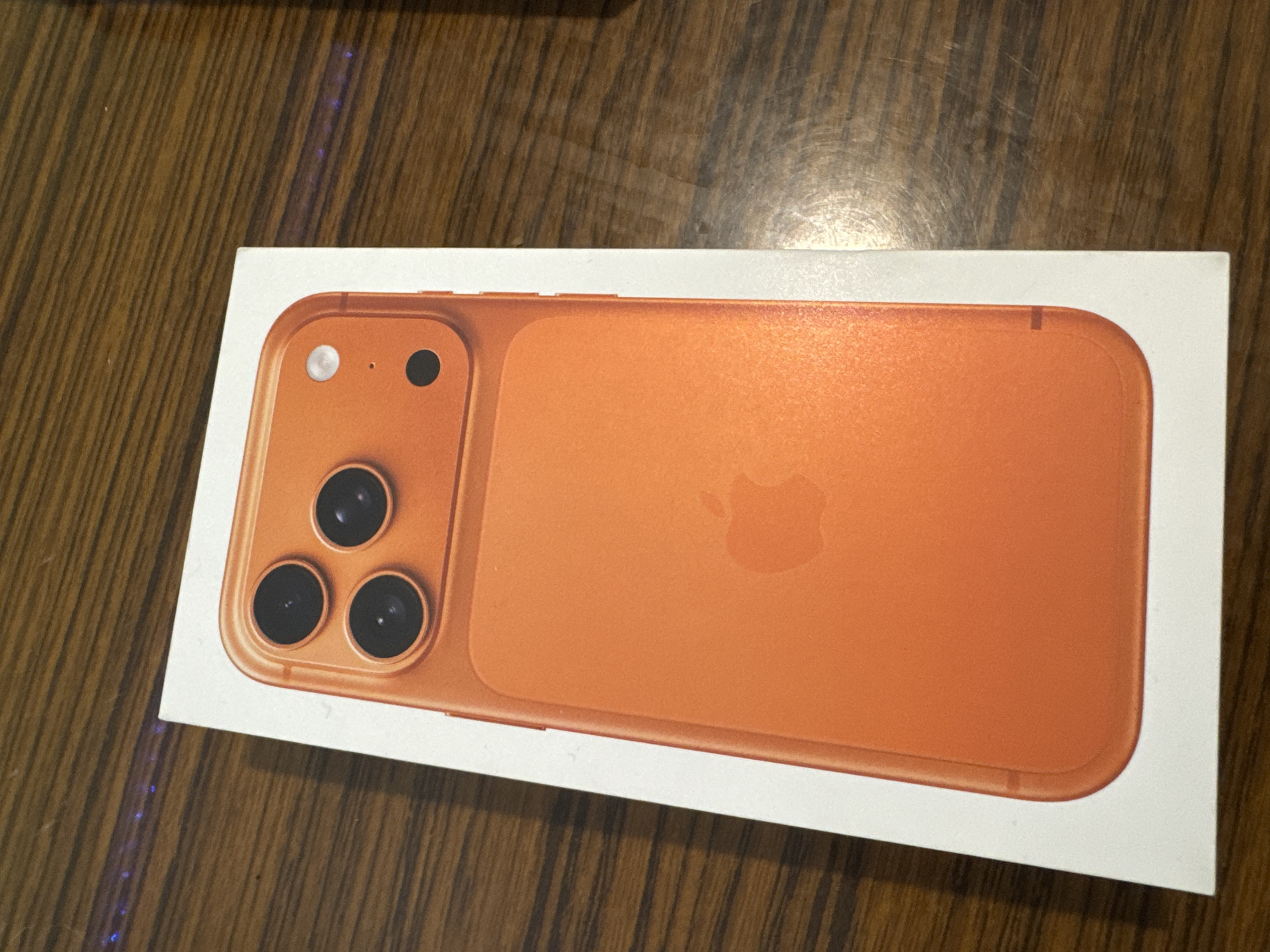 A close-up shot depicts an orange-colored representation of an iPhone on a white box, placed on a wooden surface. The phone replica features a camera array on the top left, comprising three lenses, a flash, and a microphone. An Apple logo is visible in the center of the phone's back panel. The surrounding environment consists of a wooden surface with visible grain patterns. The location of the image in the city of Yana, Bulgaria, cannot be determined from this information.