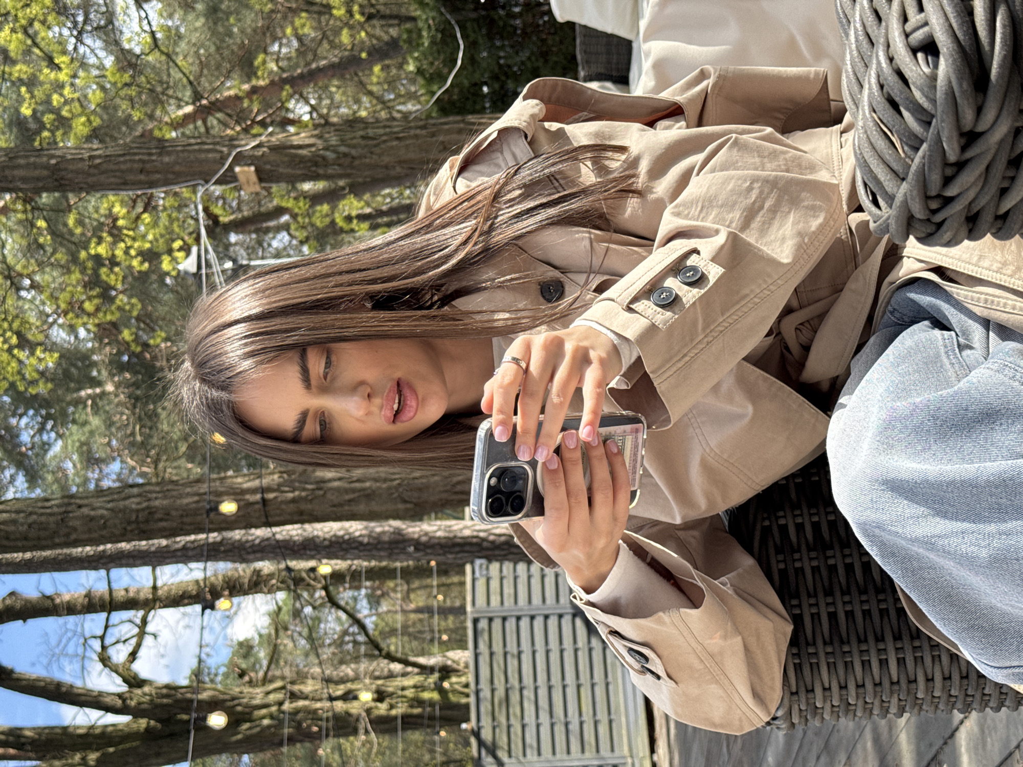 The image captures a woman seated outdoors. She has long, brown hair and is wearing a tan trench coat. The woman is holding a smartphone, possibly taking a photo or video. The scene appears to be set in a park or garden, suggested by the presence of trees with strings of lights hanging from them. A wicker-style chair is visible. The location is Konstancin-Jeziorna, Poland.