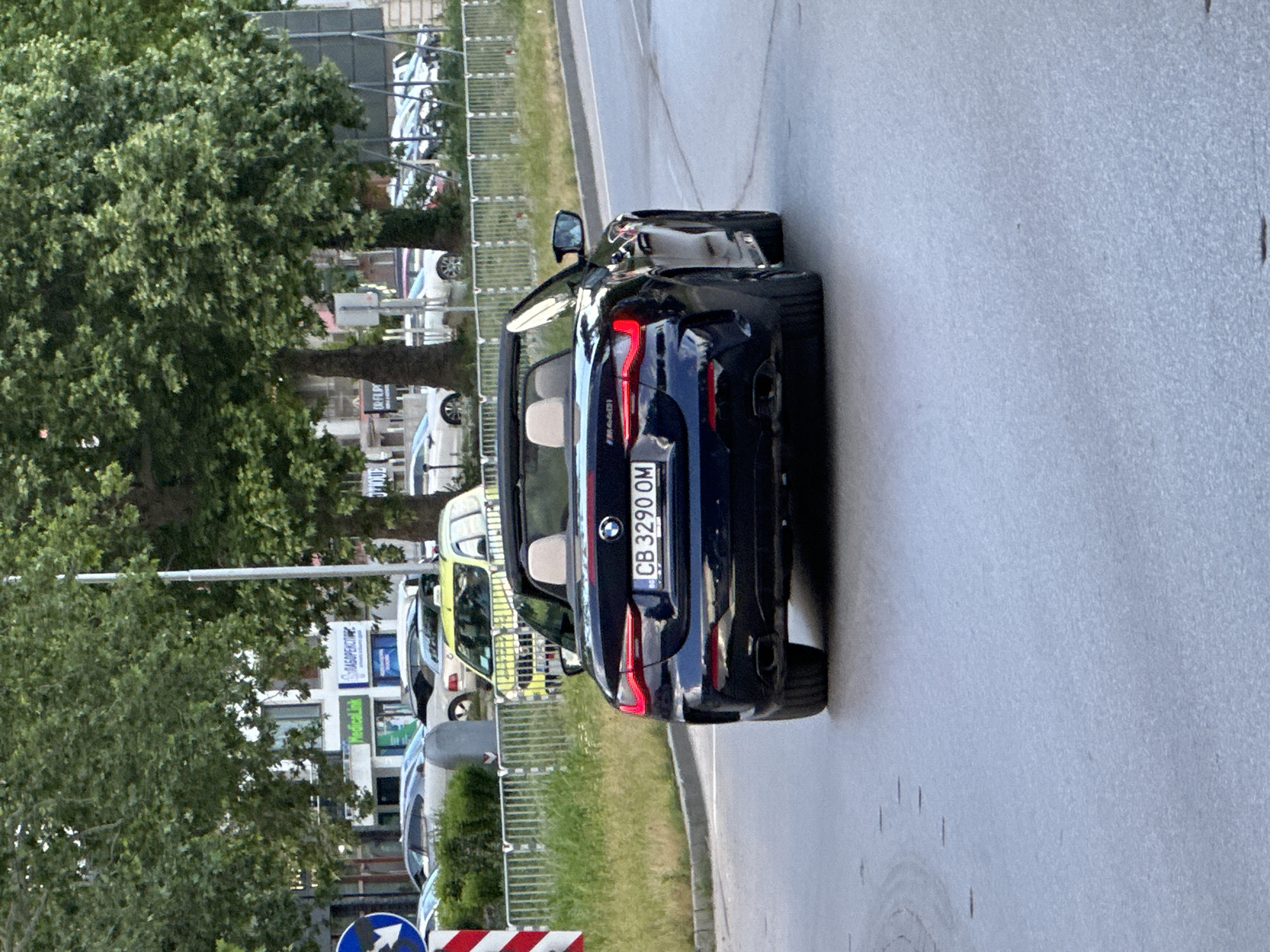 The image shows a dark blue BMW convertible with license plate CB 3290 OM parked on a road in Varna, Bulgaria. The car has its top down, revealing light-colored seats. The rear of the car is visible, including the BMW logo and tail lights. The road is asphalt and appears to be in good condition.
On the left side of the image, there is a row of parked cars, a metal fence, grass, trees, and buildings. One building has signage with the words "МЕДИКАЛСПЕКТЪР" and "Medicalink". A street sign is also visible in the lower left corner.