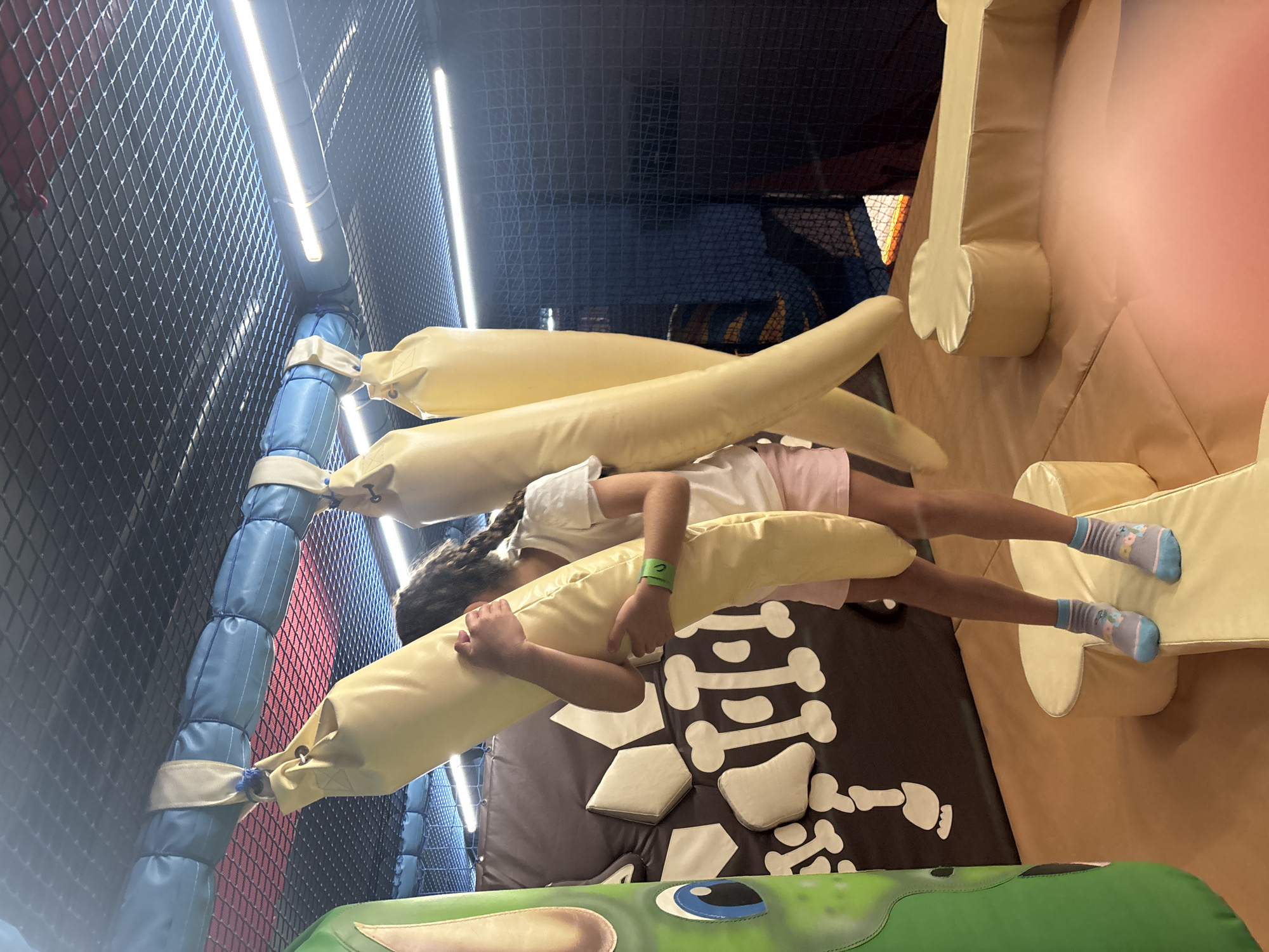 In Dubai, United Arab Emirates, a young girl with braided dark hair stands in a brightly lit indoor play area, clutching a large, cream-colored padded punching bag. She is wearing a white t-shirt, pink shorts, and striped socks with blue accents. The punching bag is one of several hanging from a padded blue bar affixed to a netted ceiling. The play area features soft, padded surfaces in shades of brown, yellow, and cream, some shaped like bones. A large, green, padded dinosaur figure is partially visible on the left side of the frame. The walls are covered with a brown padded material decorated with white bone shapes. Fluorescent lights run along the ceiling, illuminating the space.